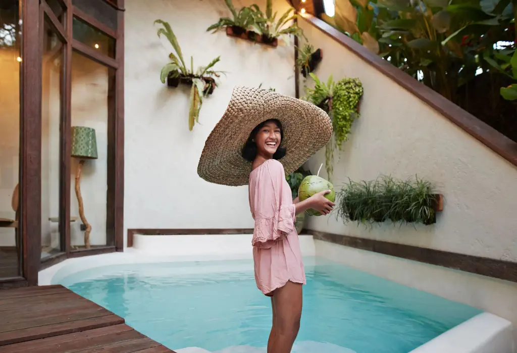 A young woman in a large straw hat smiles while holding a coconut drink in a plunge pool.