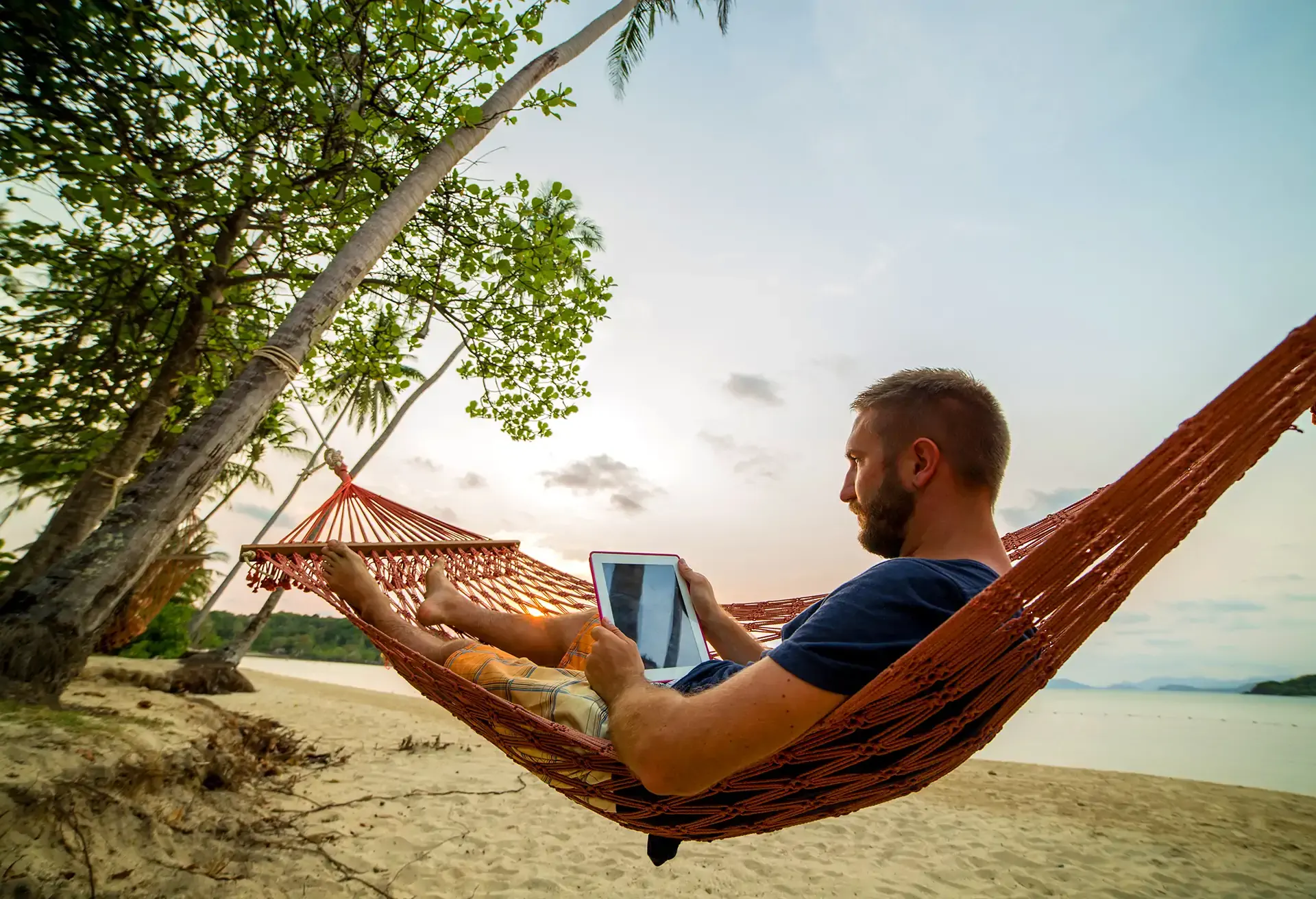 Man on a hammock reading a book from a tablet at sunset