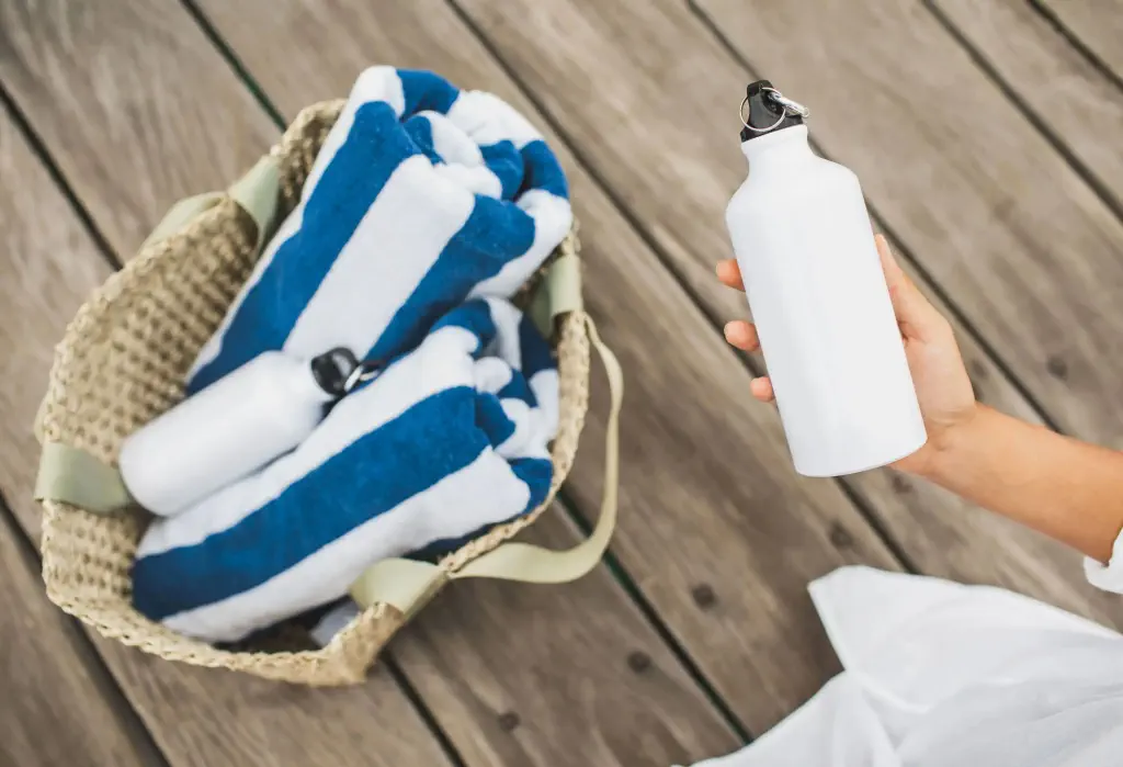 A hand holds a white water bottle near a bag with blue and white striped towels.