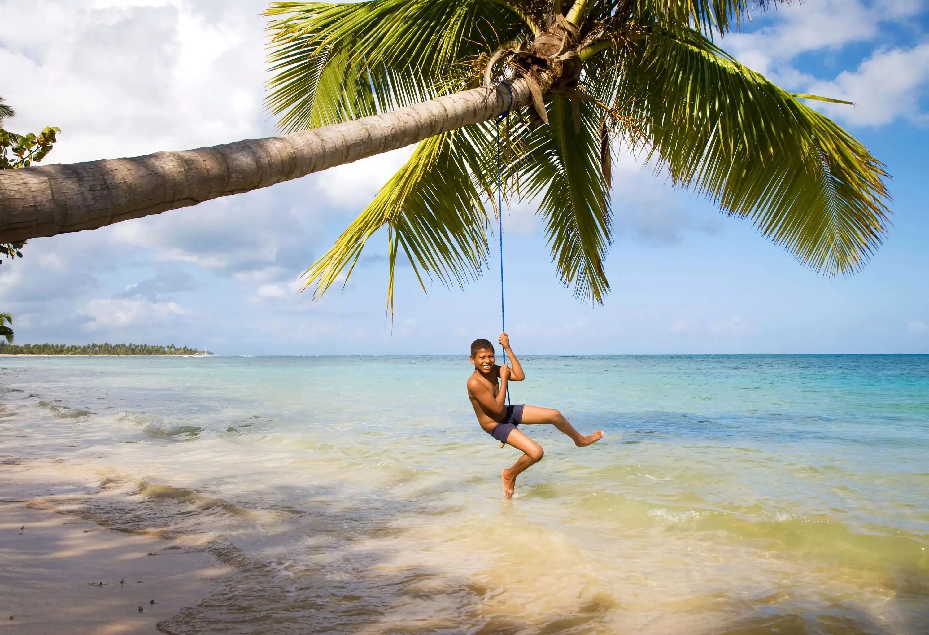 A smiling young boy in swim trunks swings from a rope tied to a leaning palm tree over shallow waters of a tropical beach.