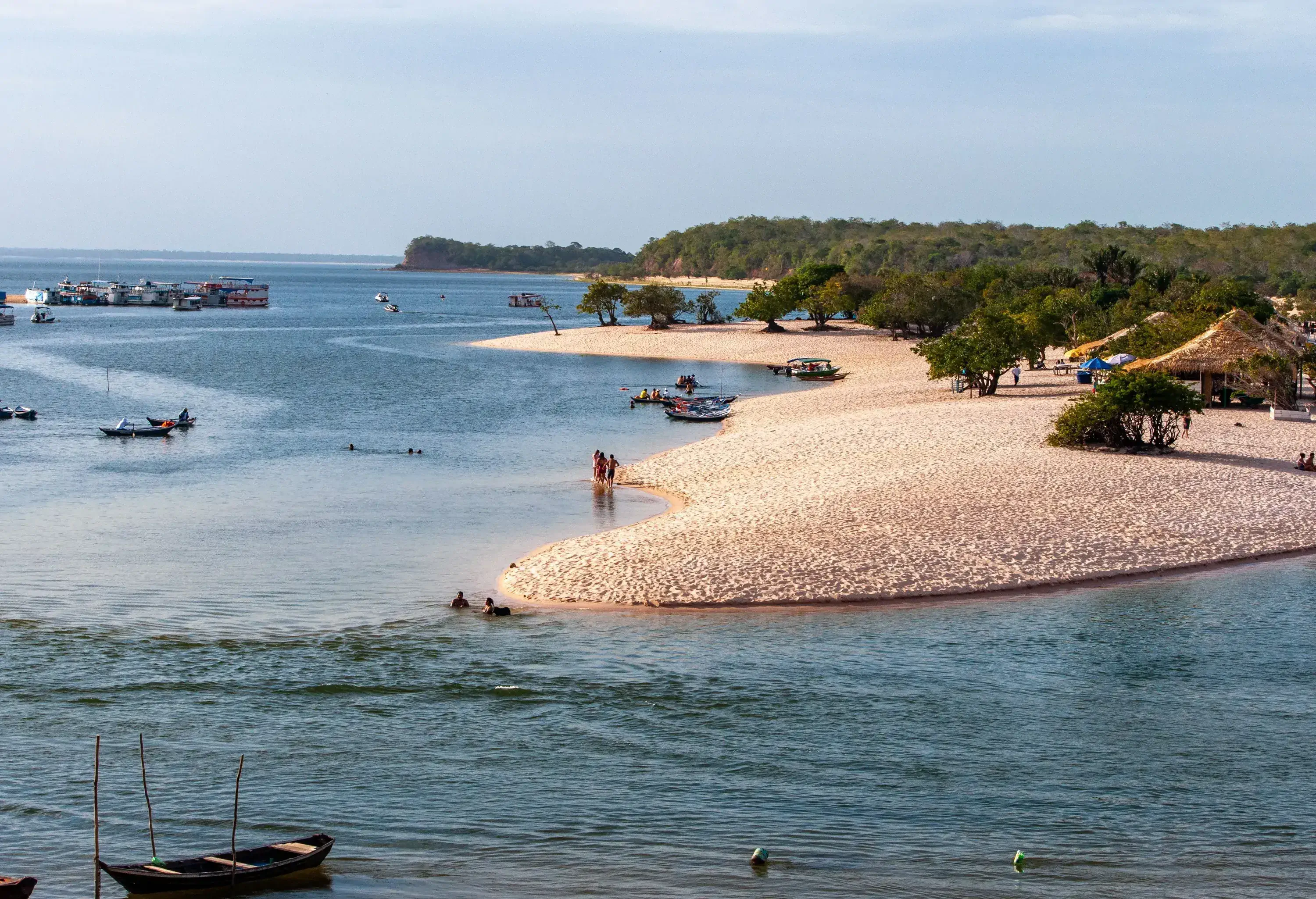 A white beach with thatched cottages amongst lush trees and anchored boats in the ocean.