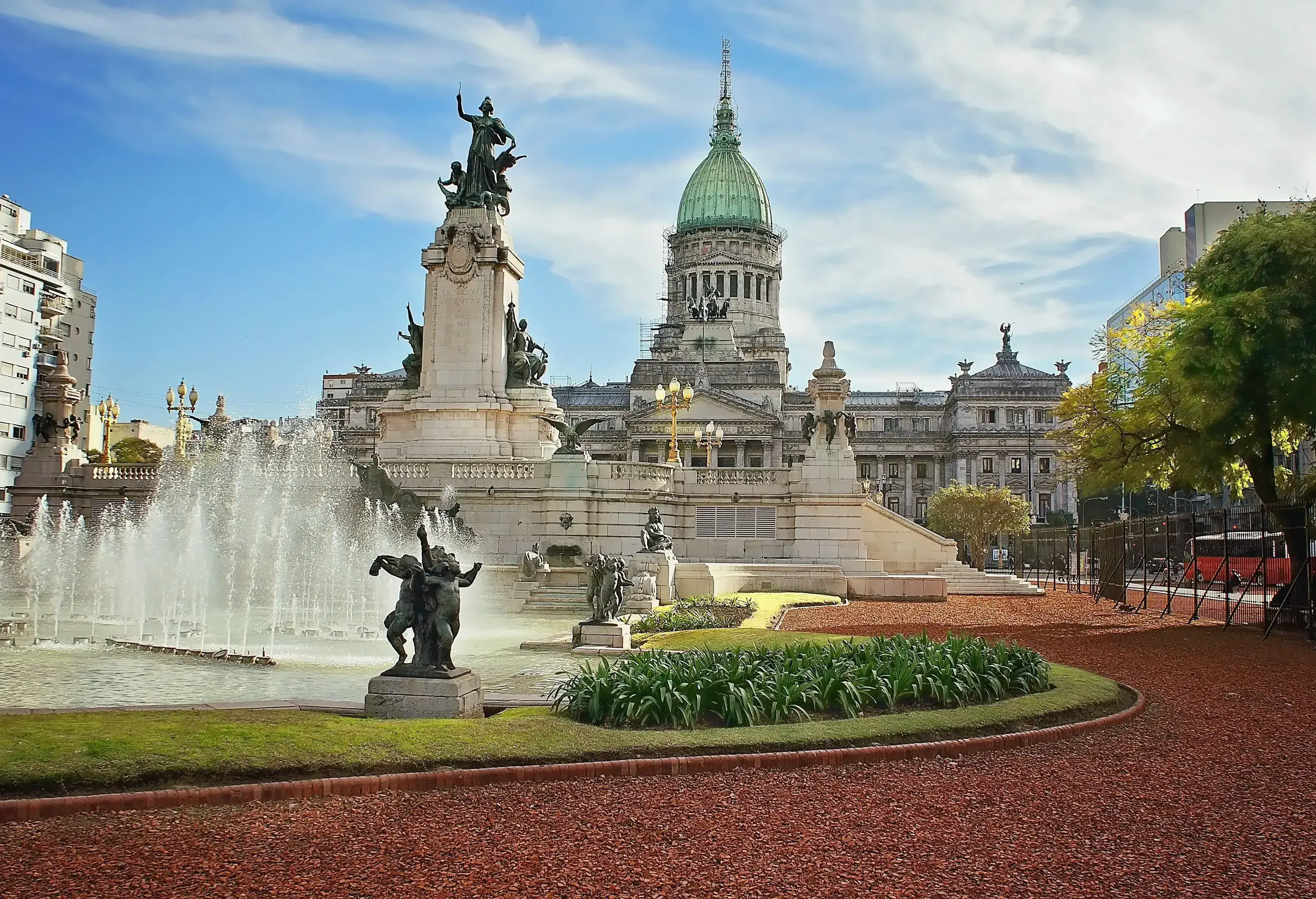 A fountain in front of a monument with bronze statues of angels and marching people, and the neoclassical white dome and exterior of Palace of the Argentine National Congress at a distance.