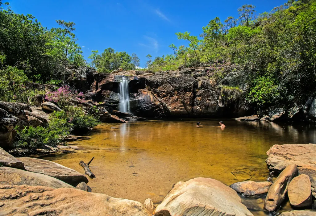 A captivating waterfall streaming down from the cliff, surrounded by lush vegetation, flowing into a clear water pool beneath where people enjoy swimming and relishing the beauty of nature's splendour.