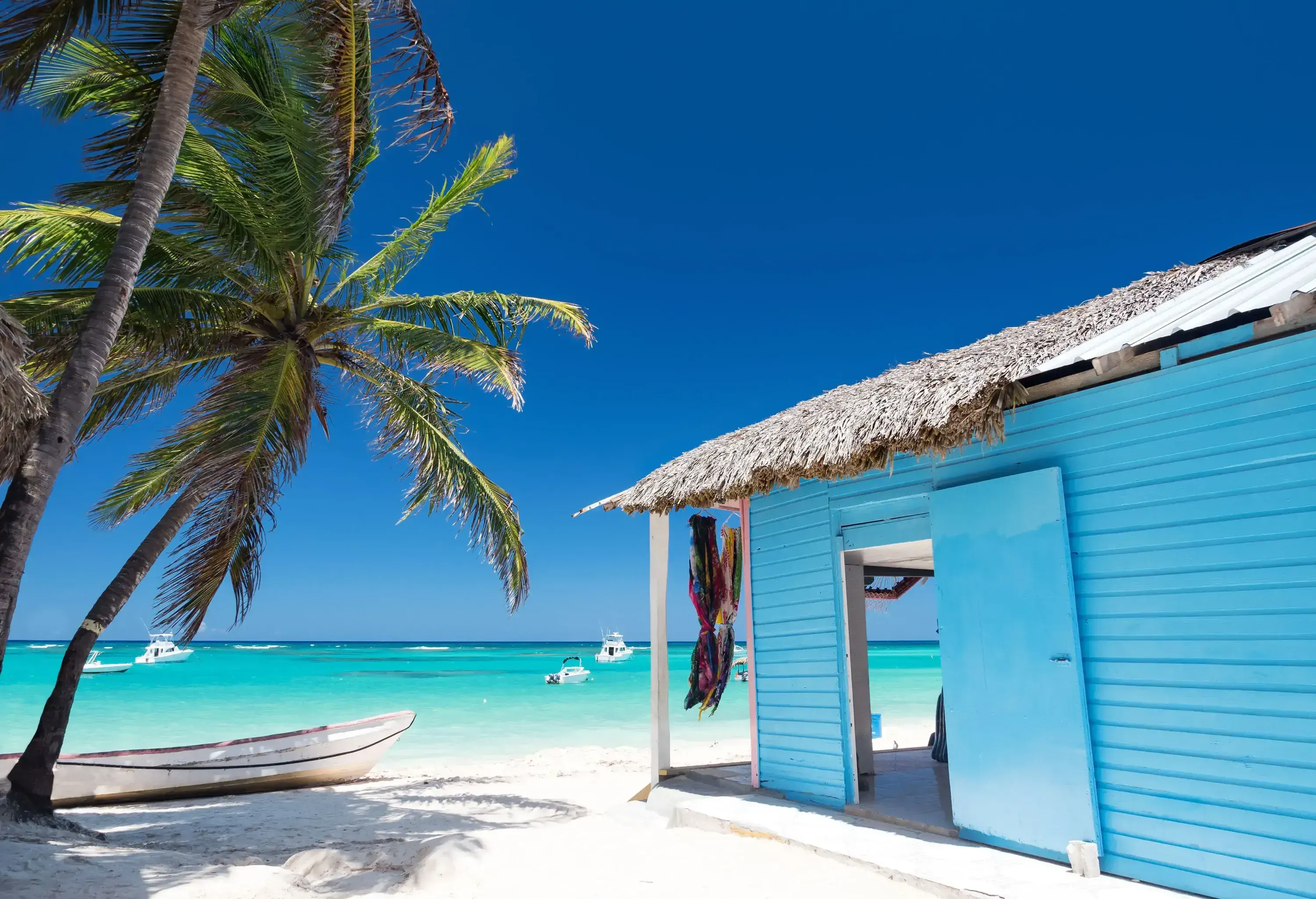 A blue wooden shed with thatched roof built on a white beach overlooking the turquoise sea.