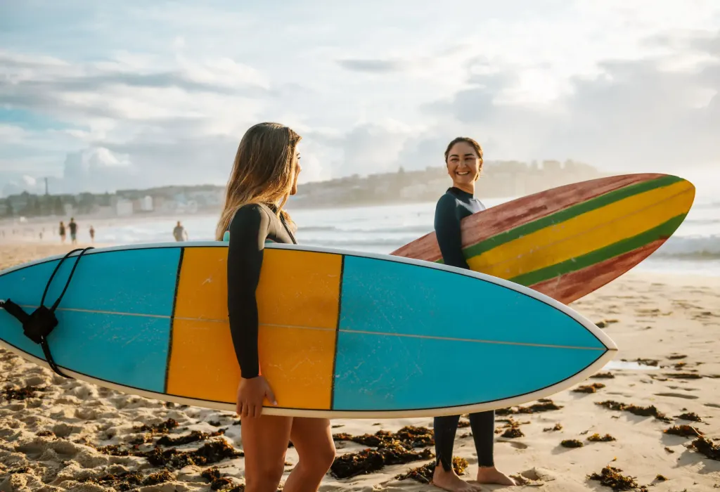 Two happy women hold their colourful surfboards as they walk on the shore towards the sea.