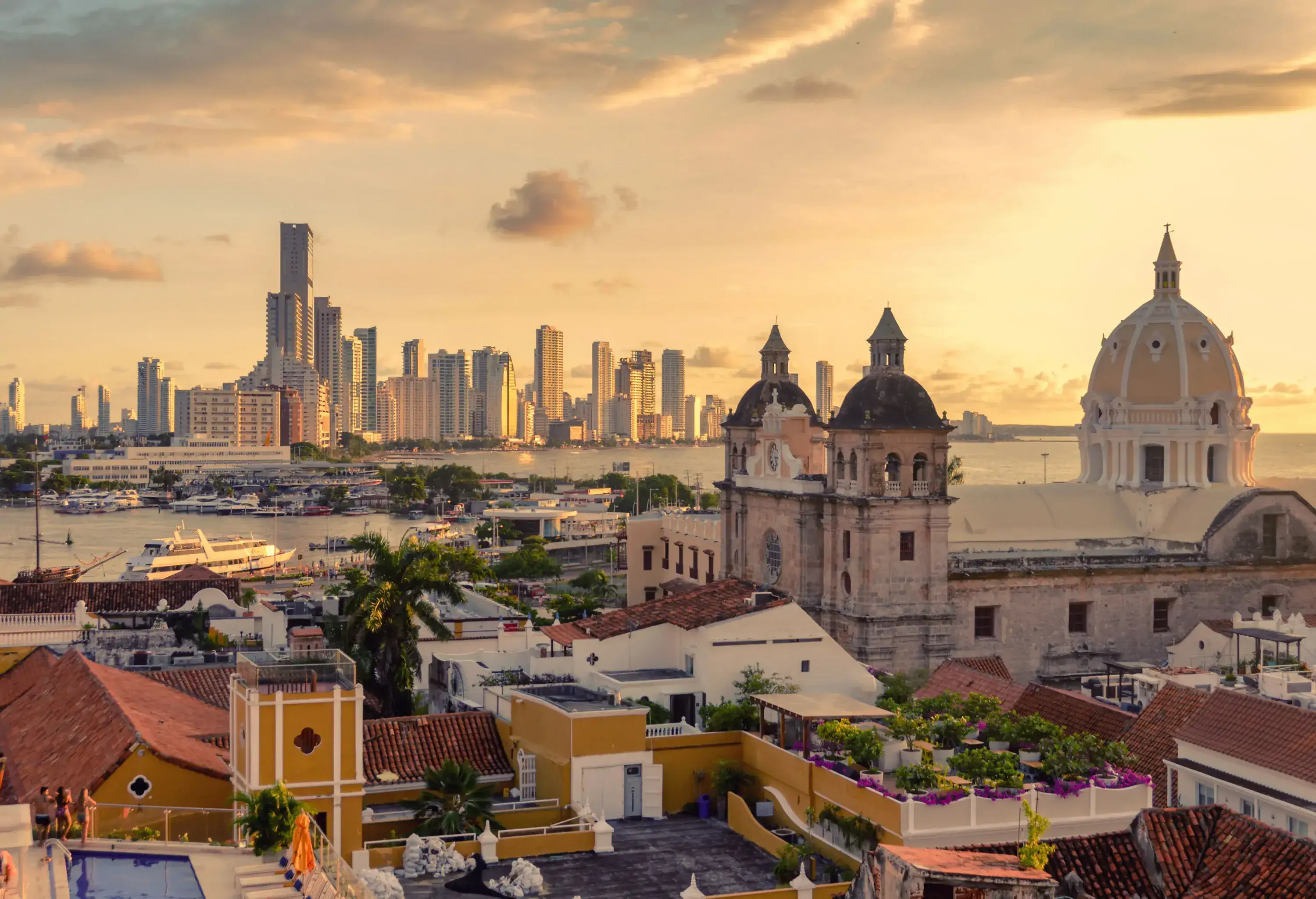 An urban area with a section of traditional buildings and grand religious edifices, facing a distant cluster of contemporary skyscrapers across a bay at sunset.