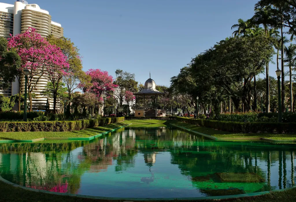 A pavilion at the edge of a green pond framed by lush green and pink trees.