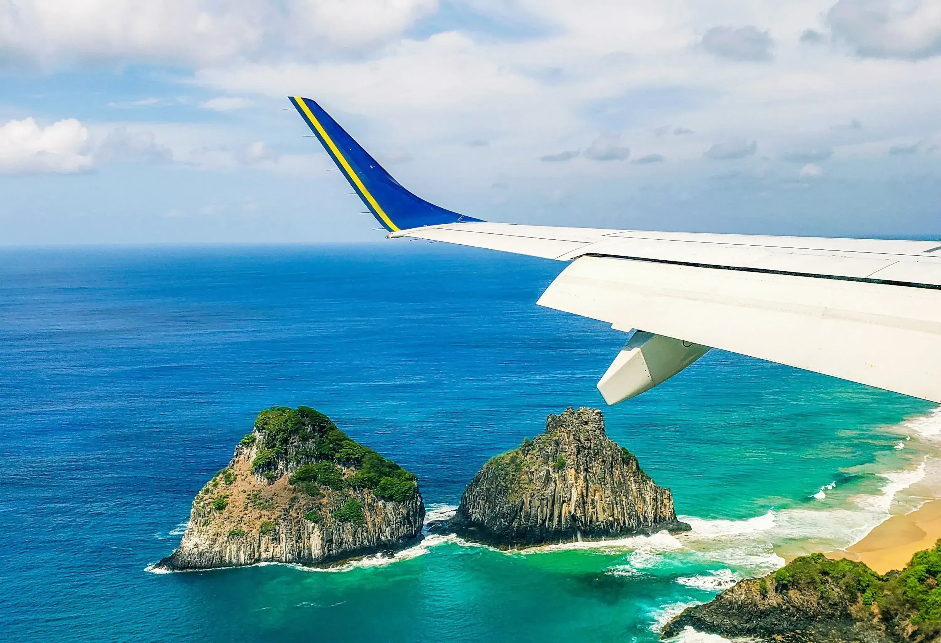 From an airplane window, the plane's wing is visible above a landscape of jutting rocks by a bay, with the vast expanse of the sea stretching into the background.