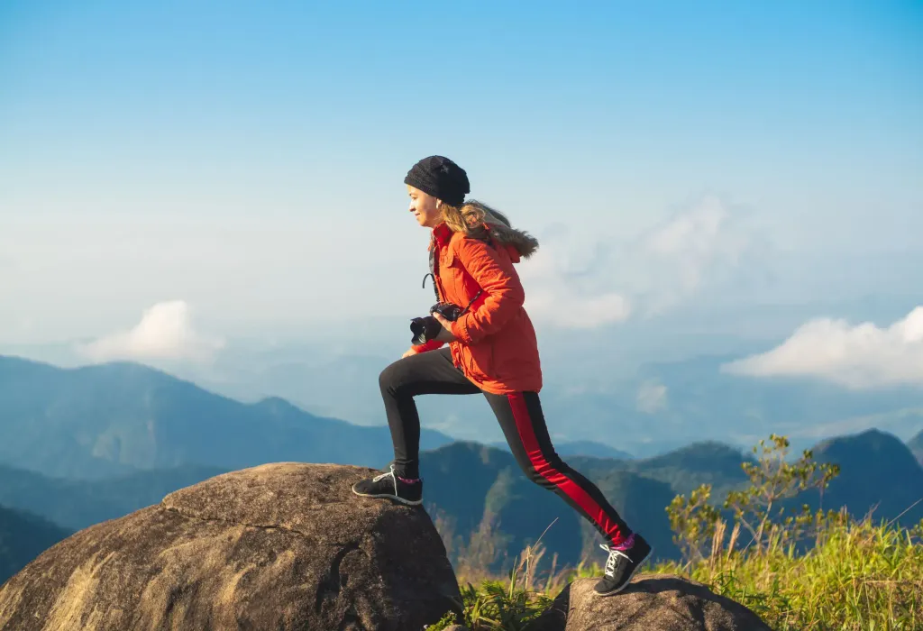 A female hiker stepping on a boulder while clutching a camera in her left hand.