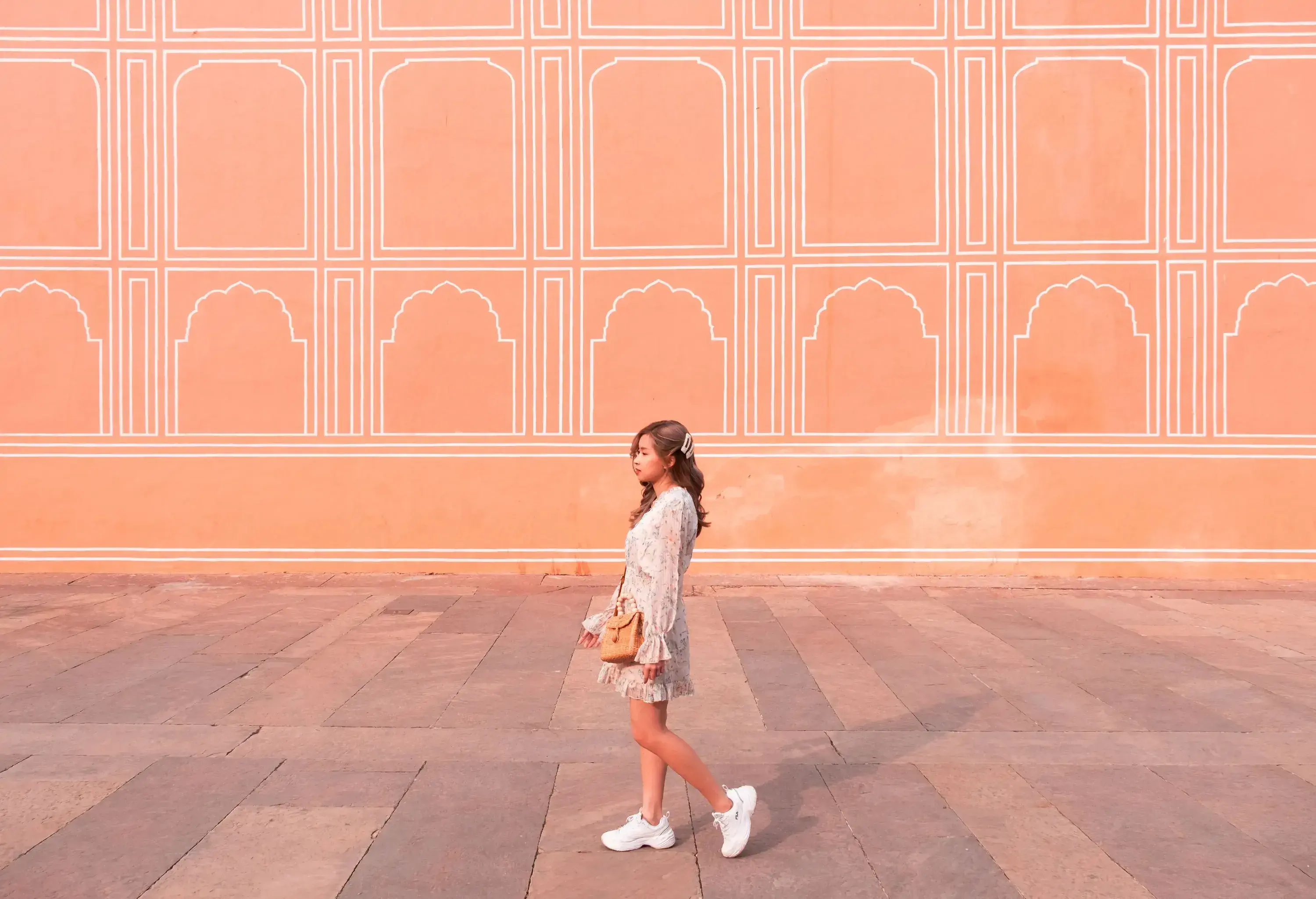 A young woman walks past a peach-colored wall adorned with white arched patterns.