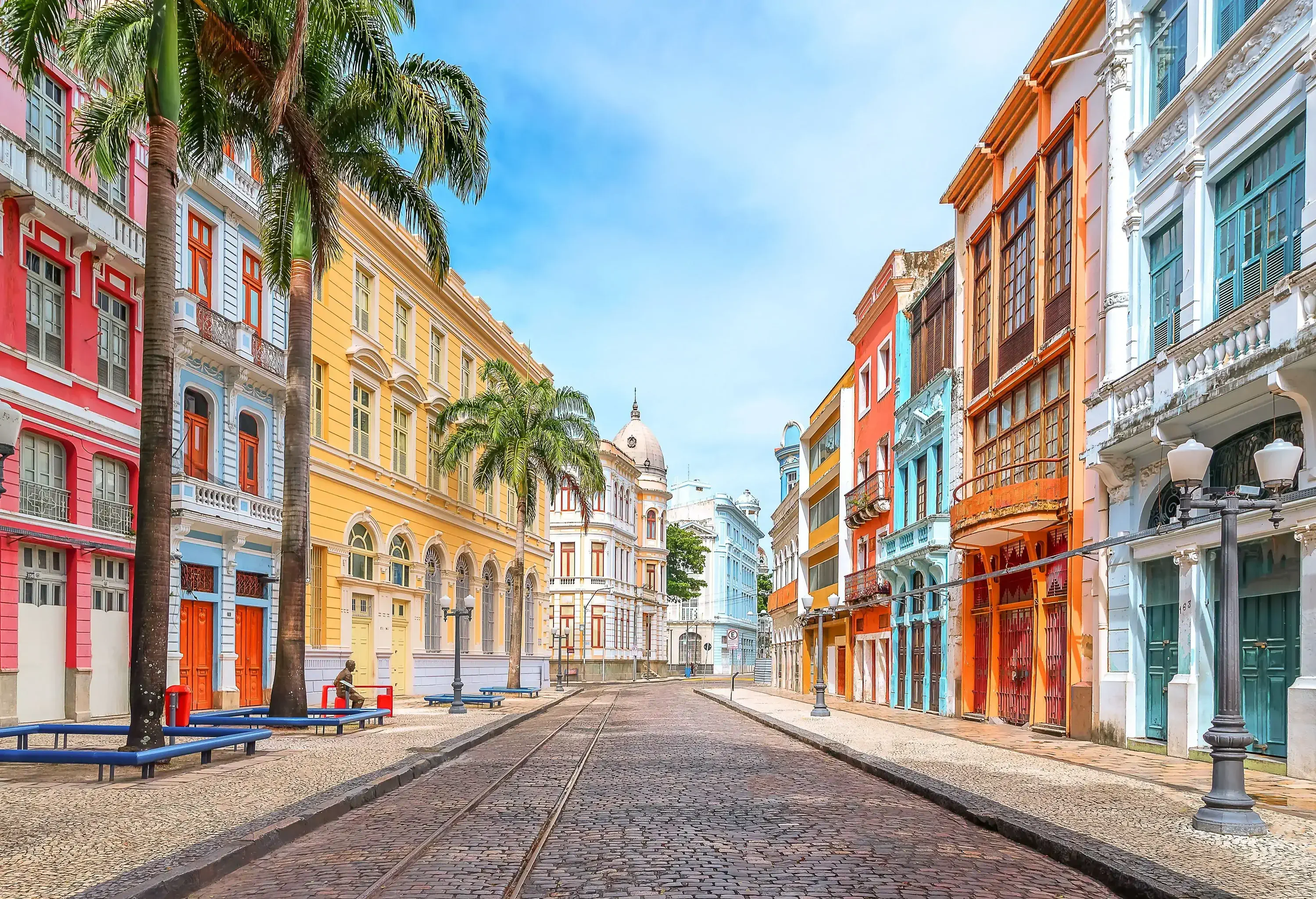 An empty cobblestone street across a row of colourful adjacent buildings with streetlamps and palm trees.