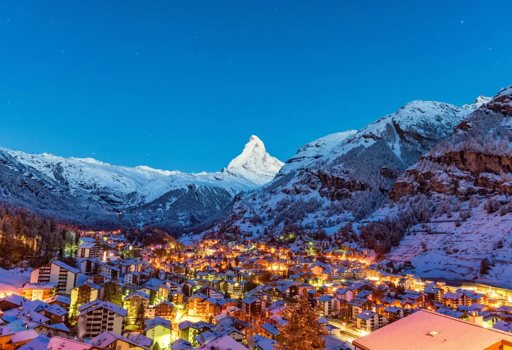 In the early morning light, a quaint town with brightly lit houses comes to life, nestled amidst the majestic snow-covered mountains.