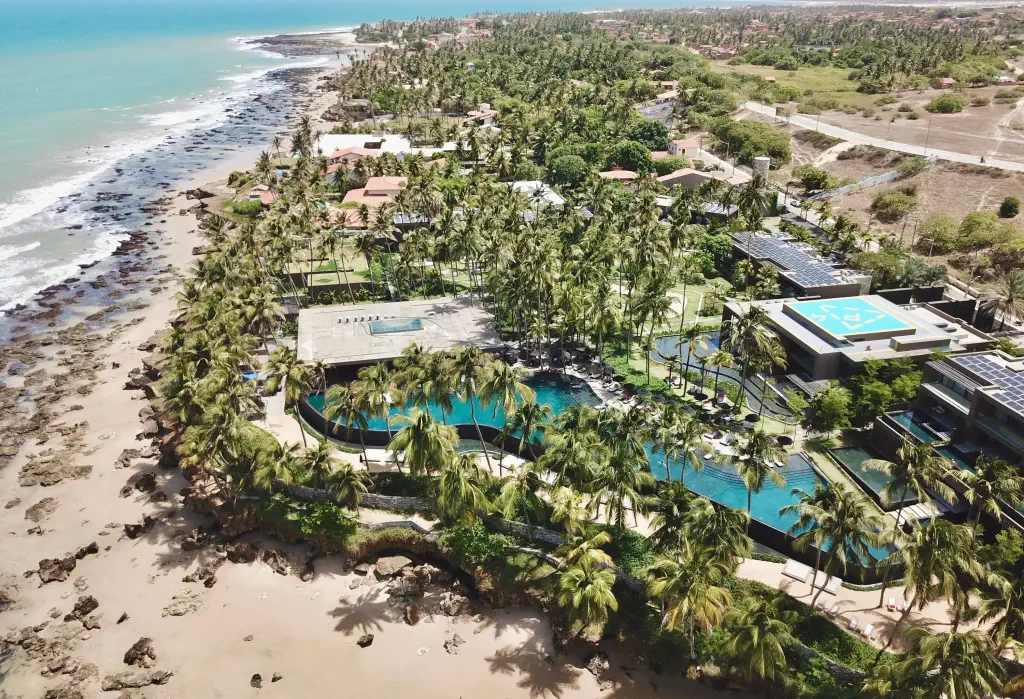 A resort with a big swimming pool surrounded by a grove of palm trees along a beach with coastal rocks.