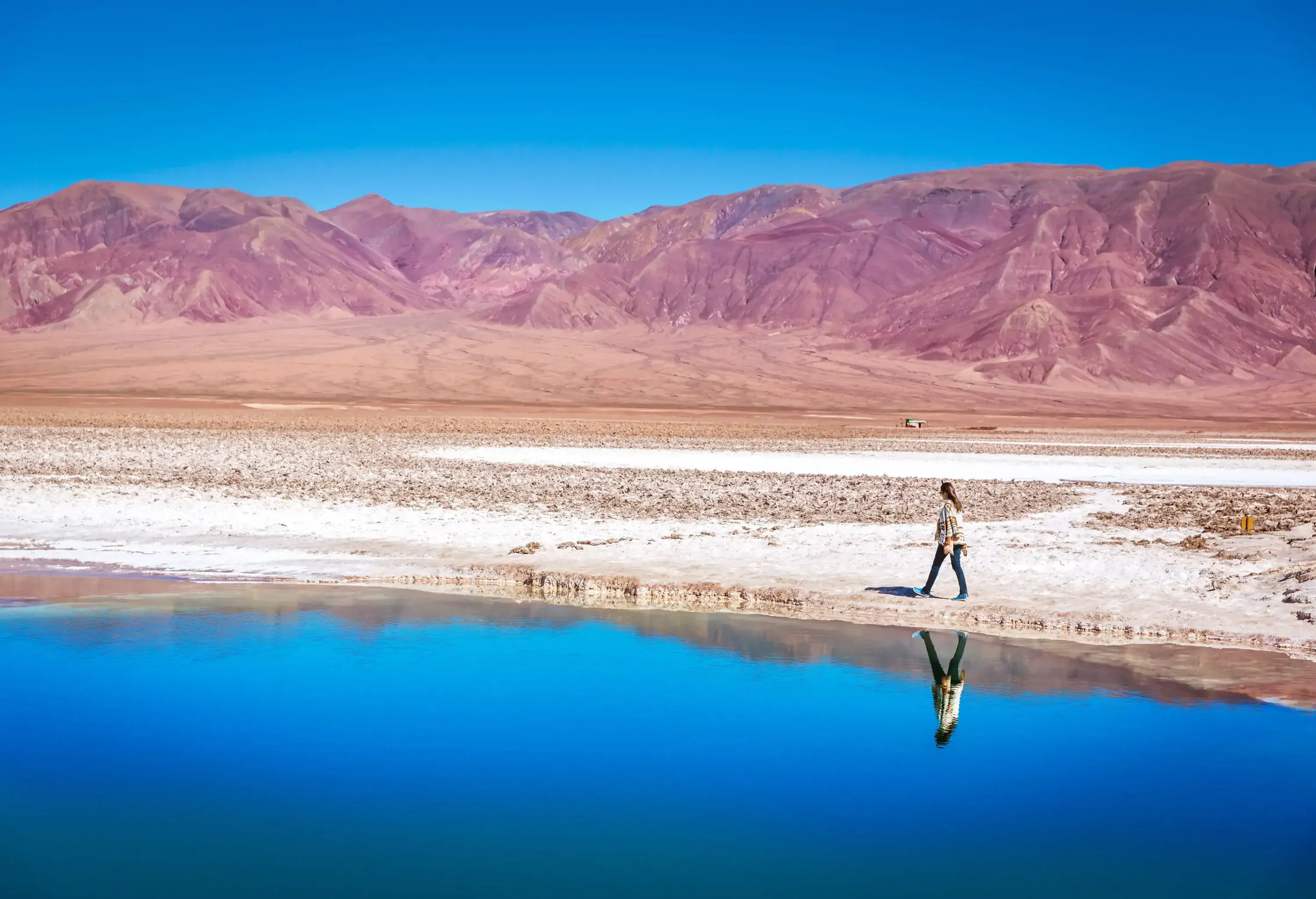 A woman walking along a salt-crusted shore of a lake in a mountainous desert.