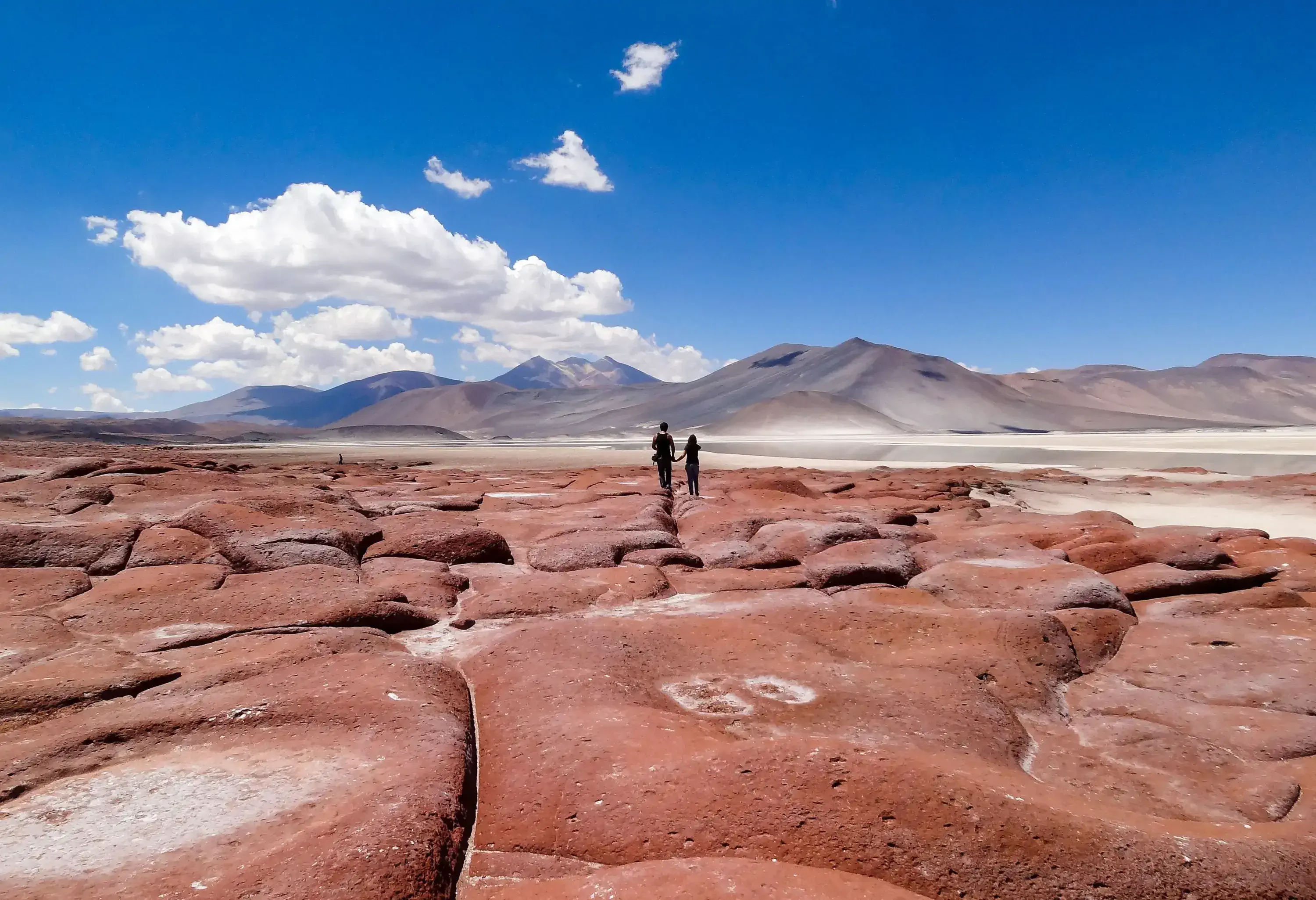 A man and a woman holding hands as they stand on a red bedrock in the foothills of a sandy mountain range.