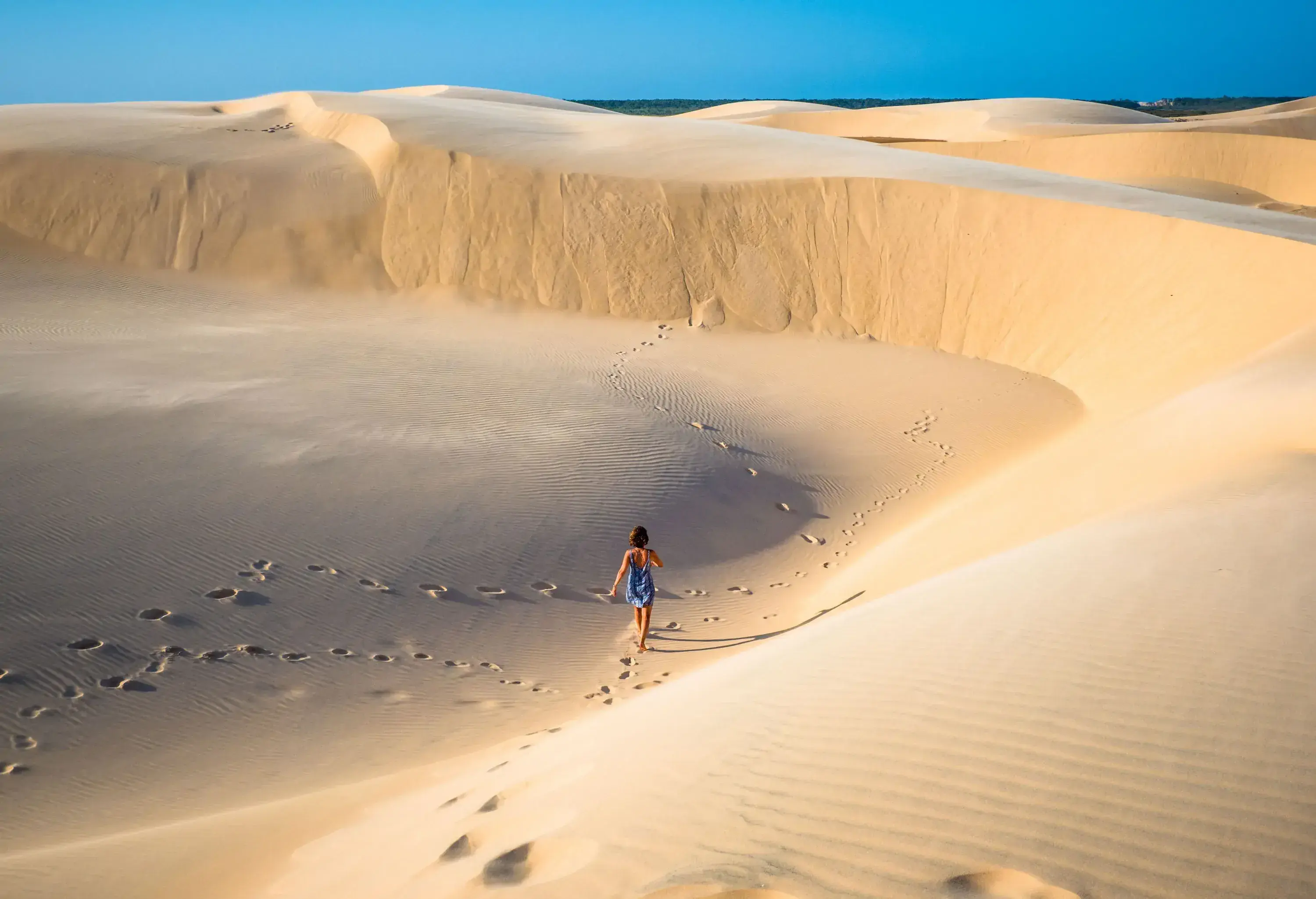 A woman wandering across a fine sand dune with traces of footprints behind her.