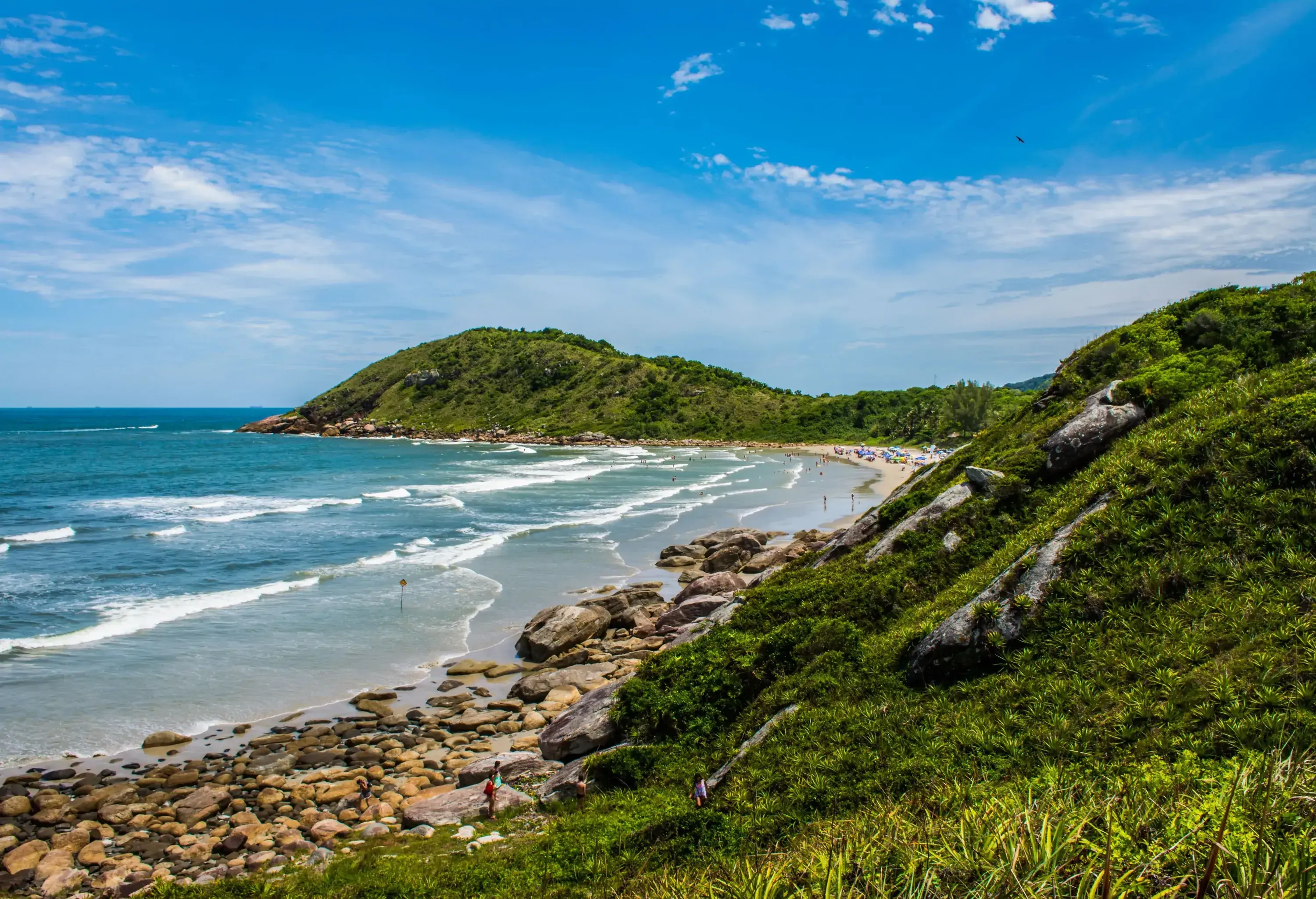 A lush island with a rocky shore by the wavy turquoise sea against the cloudy blue sky.
