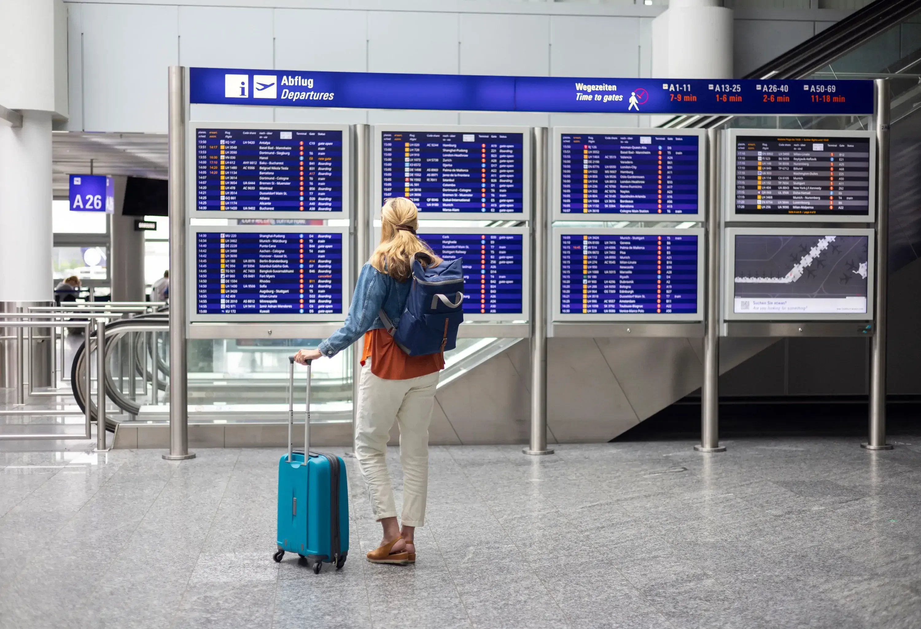 a woman standing with her suitcase in front of the airport departure boards. She is looking at the times for the flights on the boards while wearing casual clothing and carrying a backpack.
