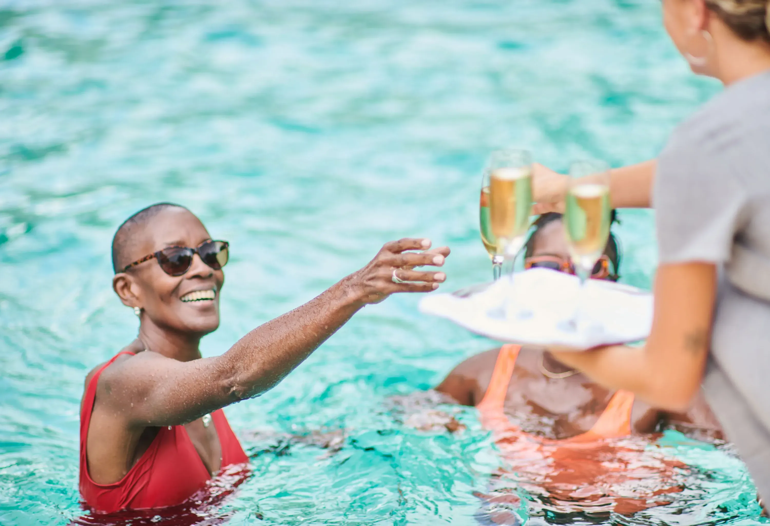 A person in a pool receives a drink from another who holds a small serving tray with sparkling wine glasses.