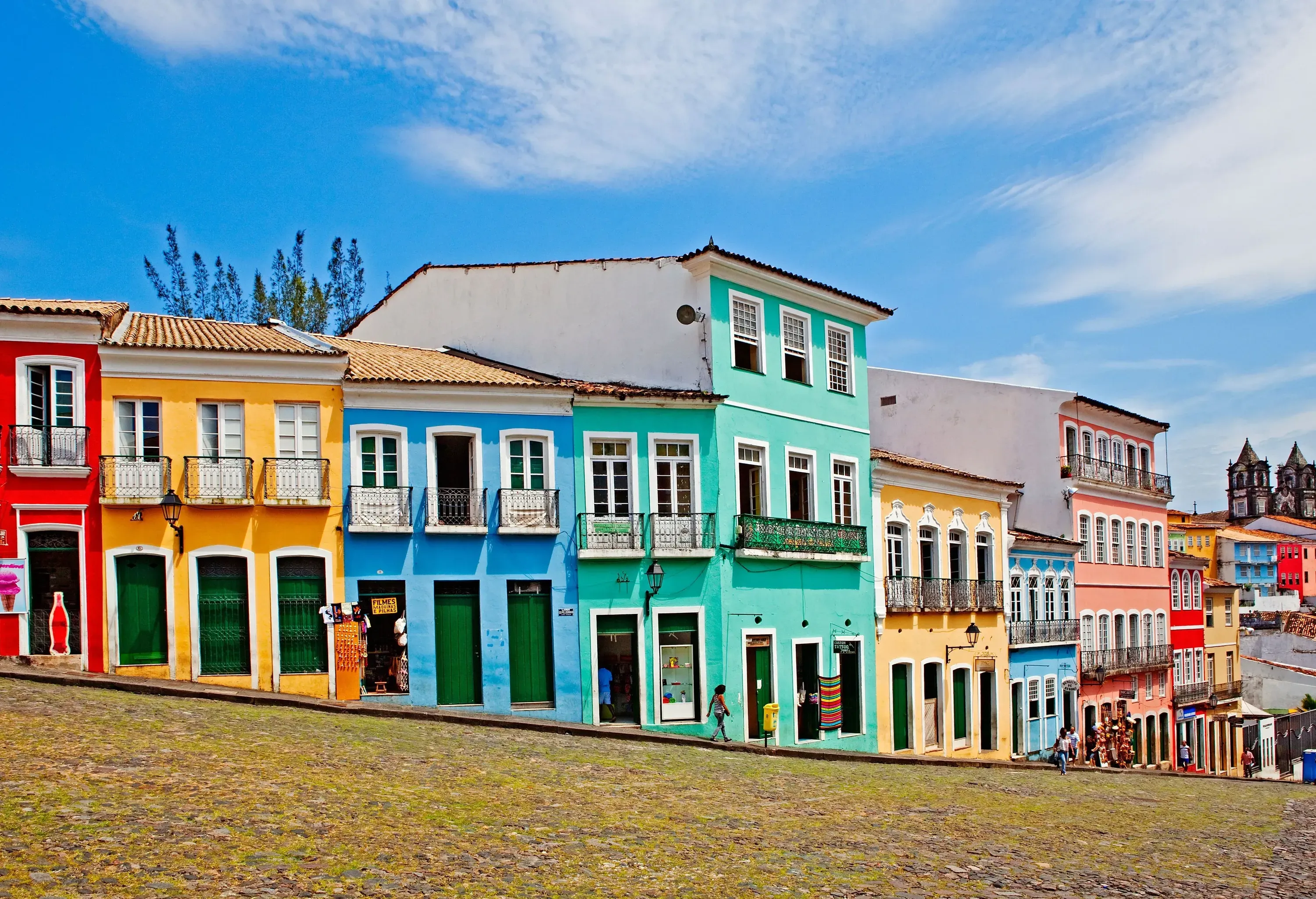 A street descending on a slope, lined with colourful wall-to-wall buildings, peppered with a few pedestrians going about their day.