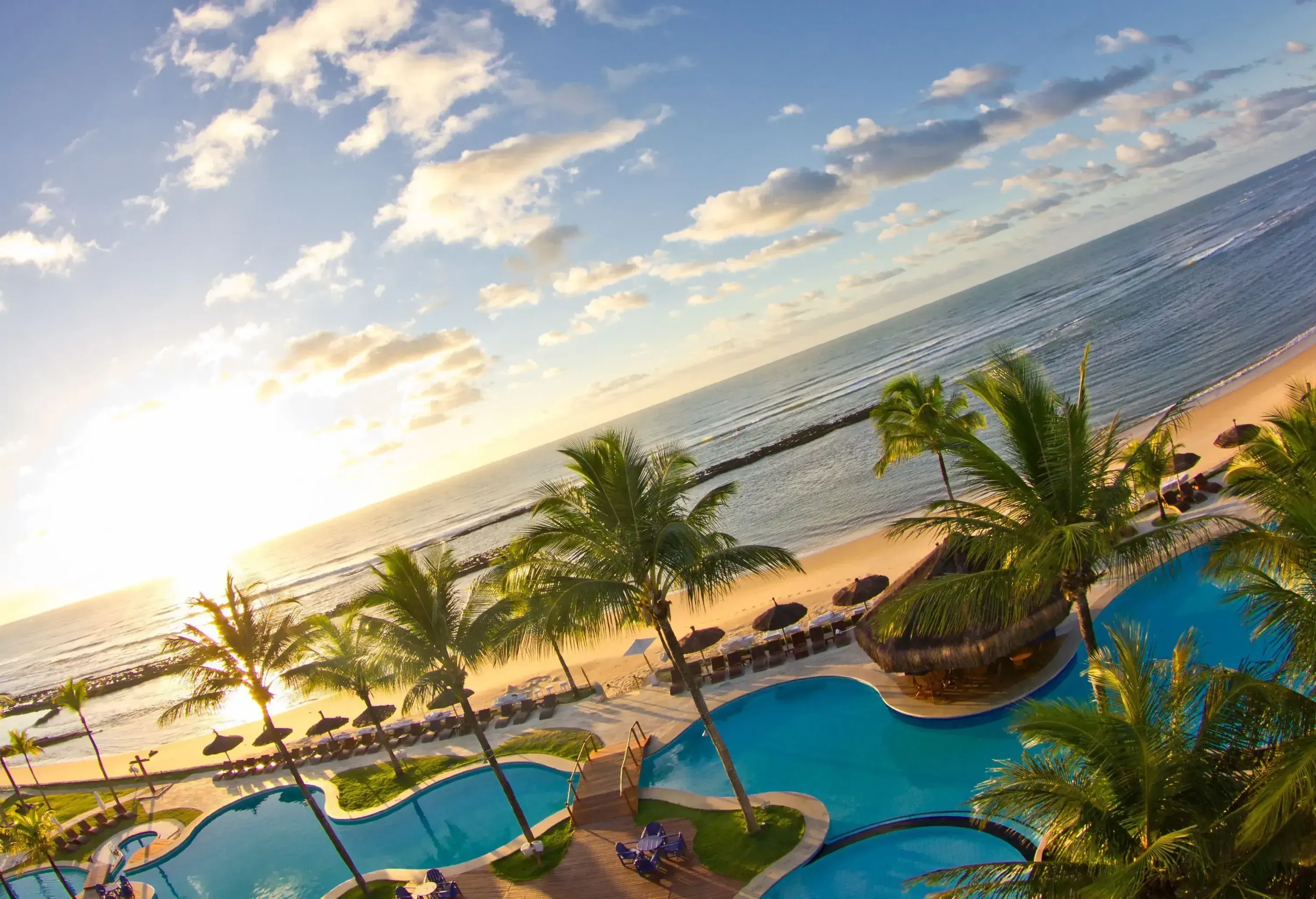 A wavy-patterned swimming pool dotted with palm trees, next to a beach lined with beach beds and parasols, with the sea in the background.