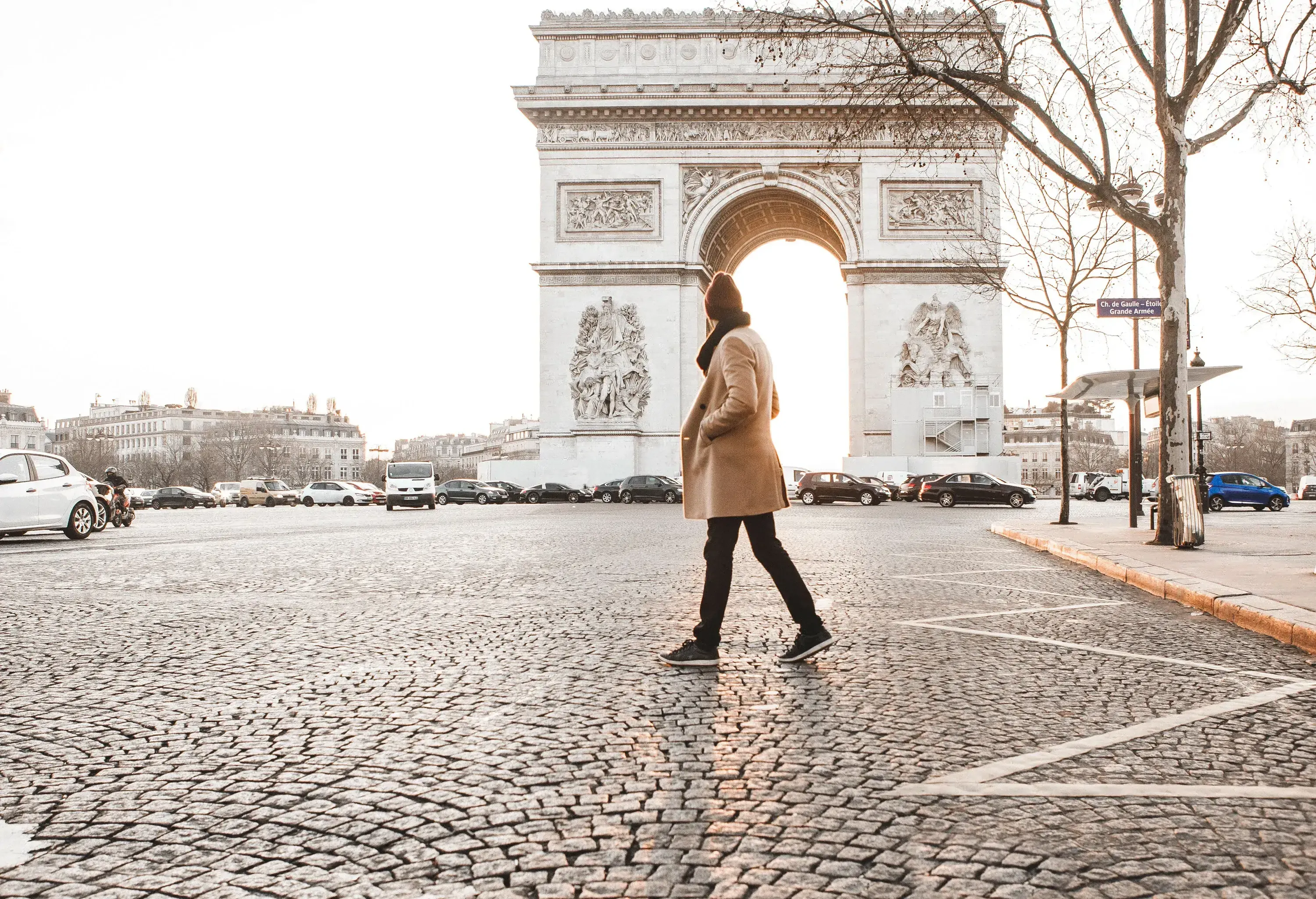 A man in a winter coat walking on the cobblestone street in front of the Arc de Triomphe.