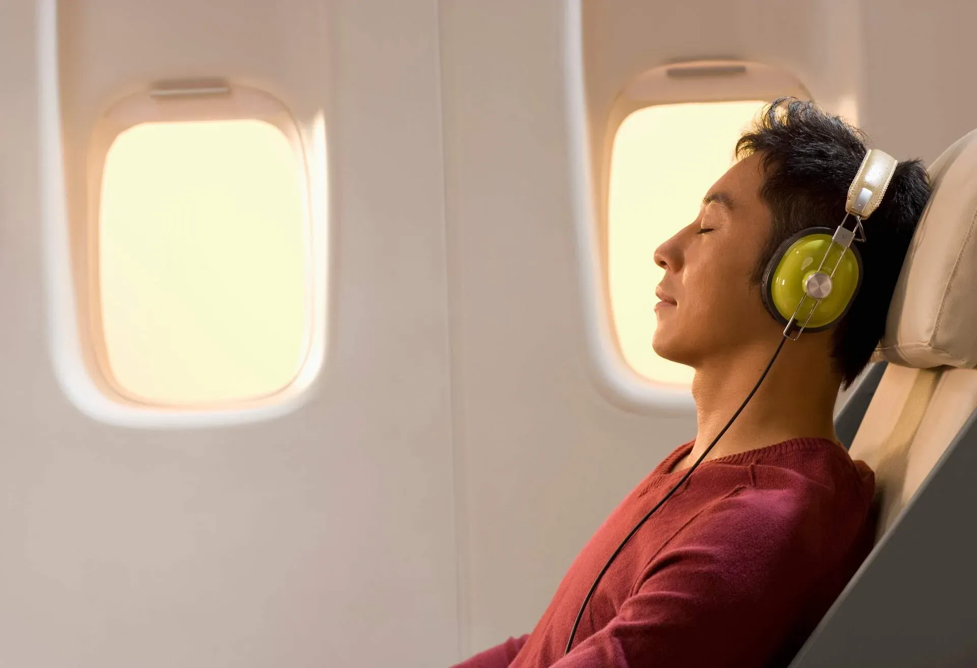 A male passenger in a window seat falling into slumber with headphones over his ears during a flight.