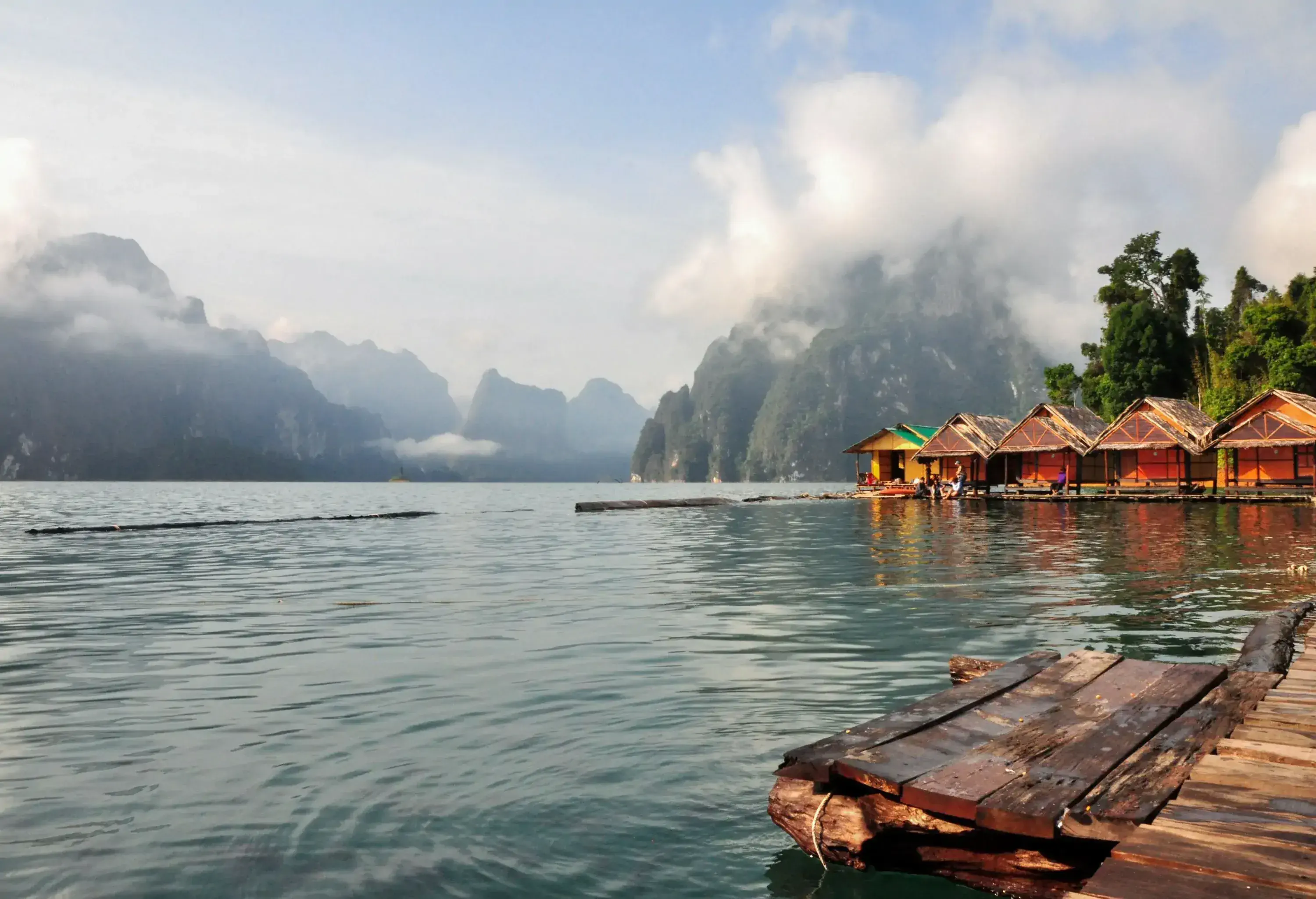 A tranquil lake lined with wooden houses and forested mountains surrounded by clouds.