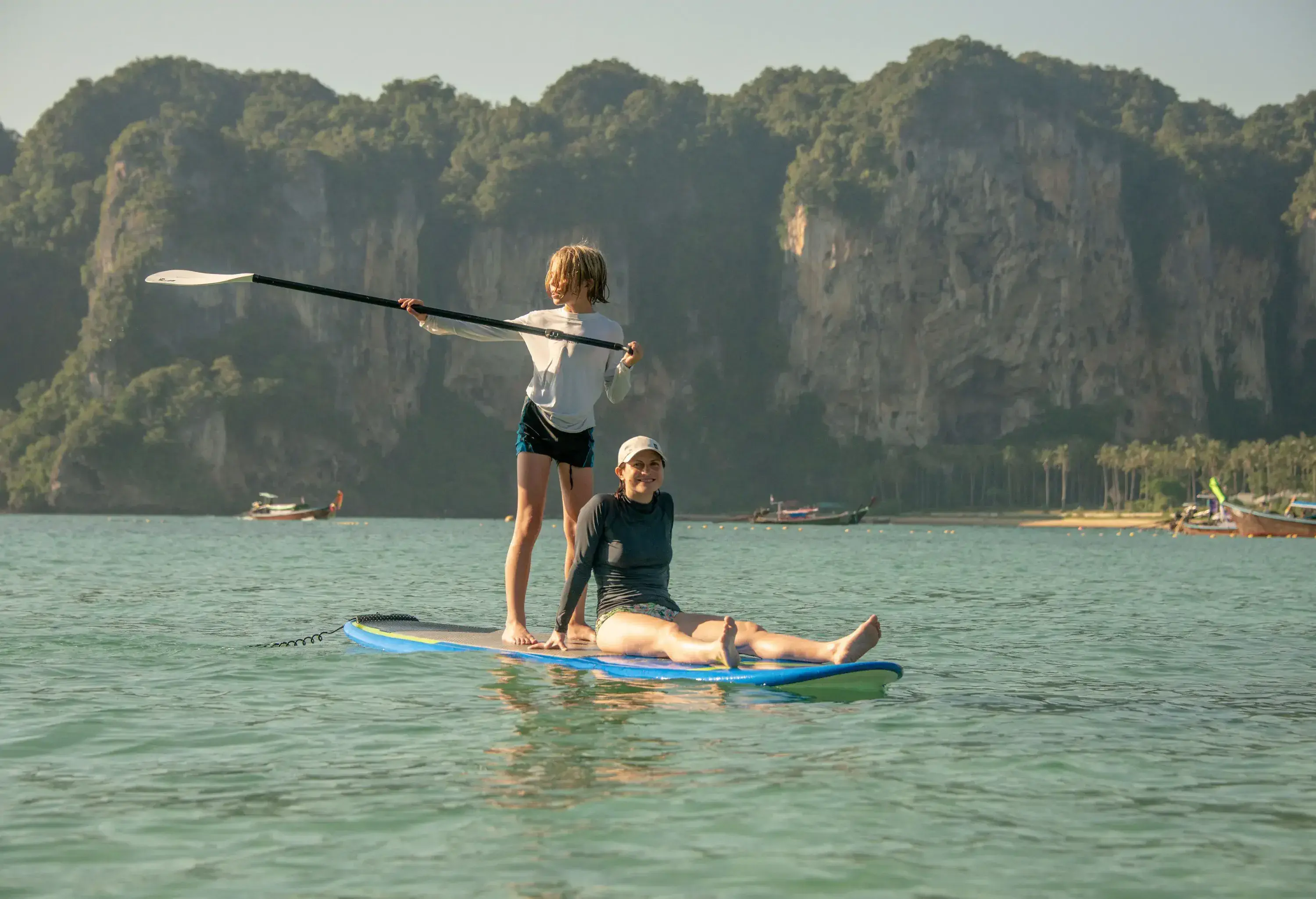 A teenage boy and his mother are seen paddling on a surfboard against the backdrop of a coastal cliff.