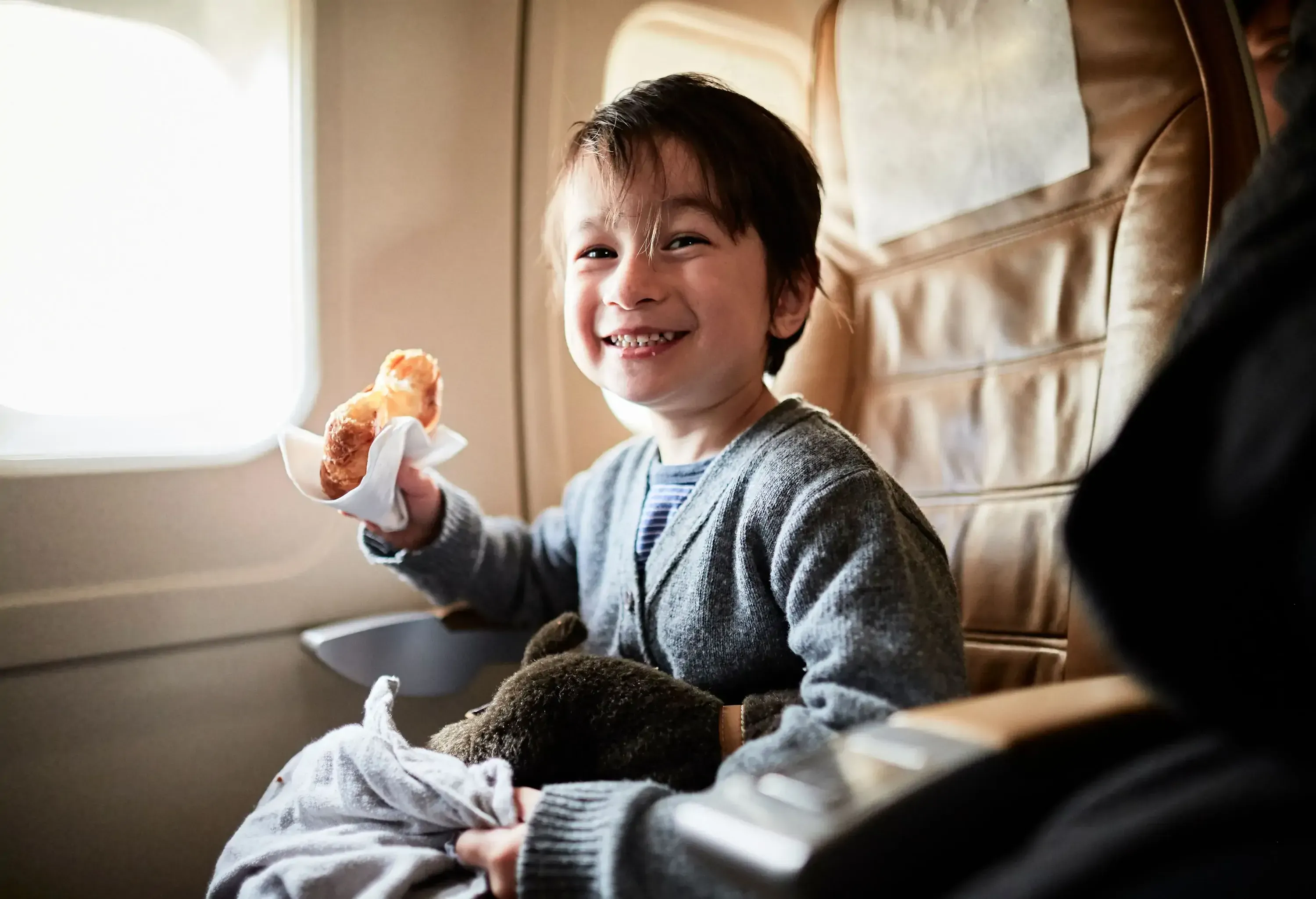 A young boy holding a snack smiles happily while sitting in an airplane seat.