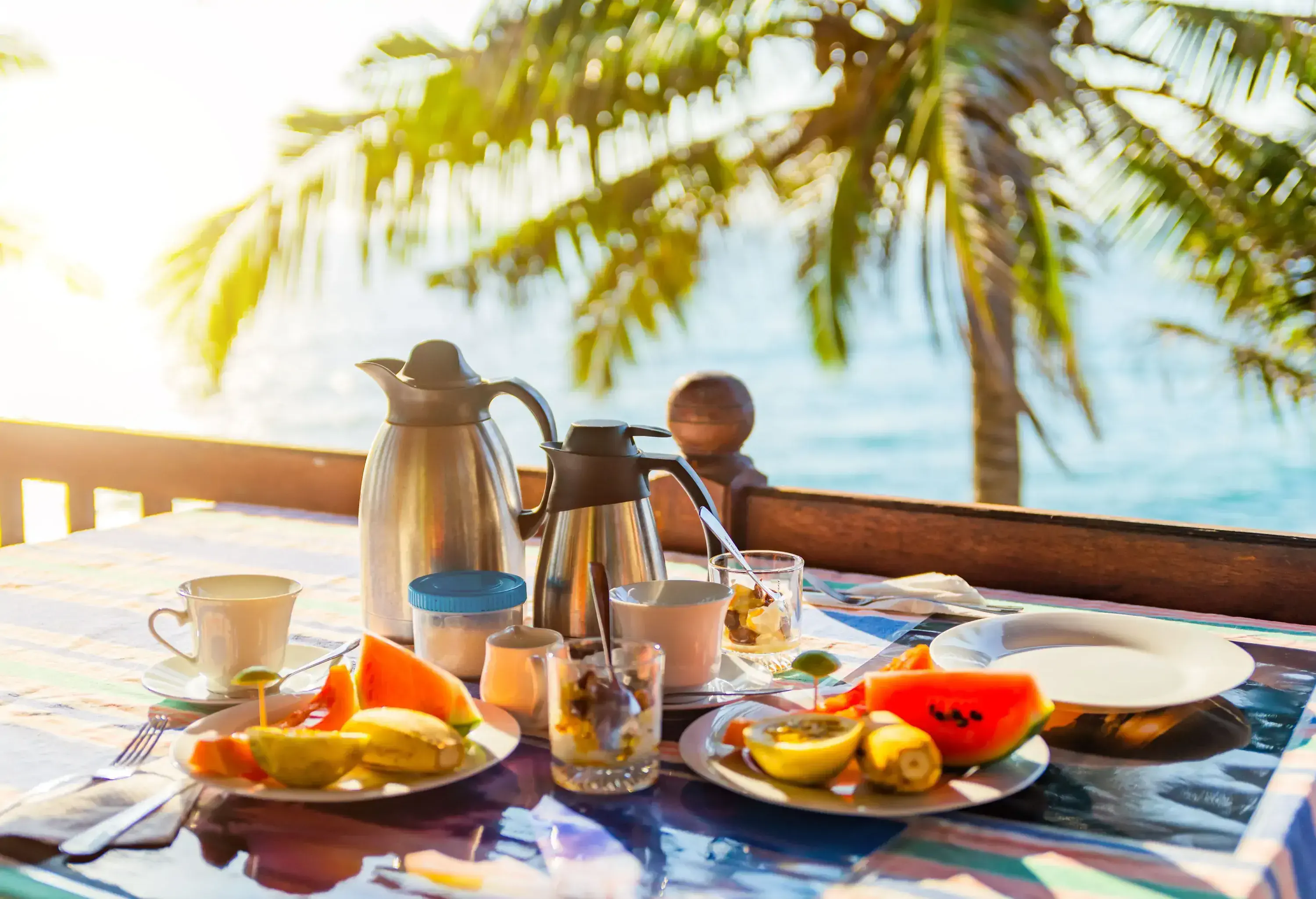 A healthy tropical breakfast with fresh fruits and hot beverages is served on a table overlooking the sea.