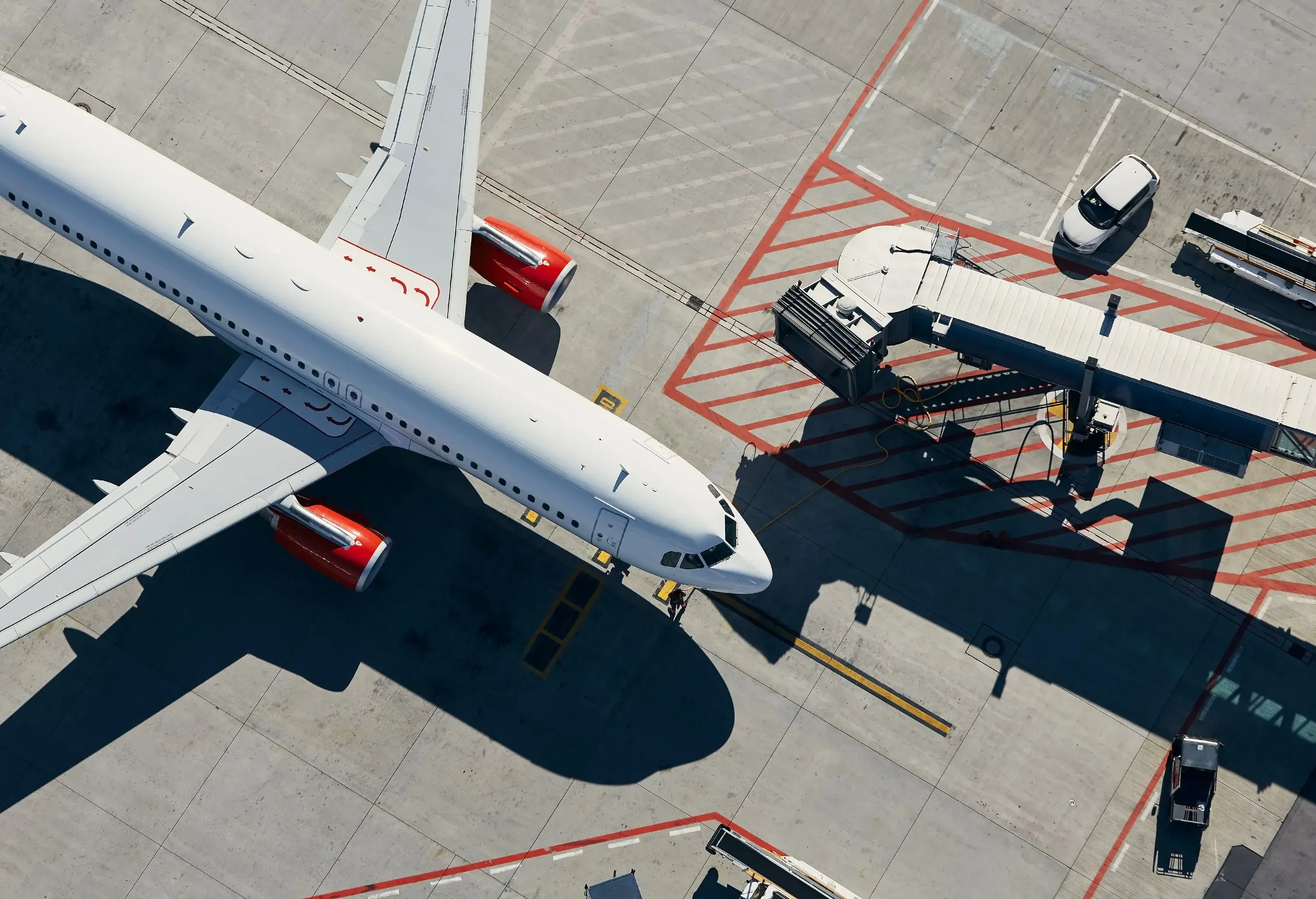 An aerial view of a large white airplane, along with a jet bridge, ground vehicles, and painted lines on the concrete.