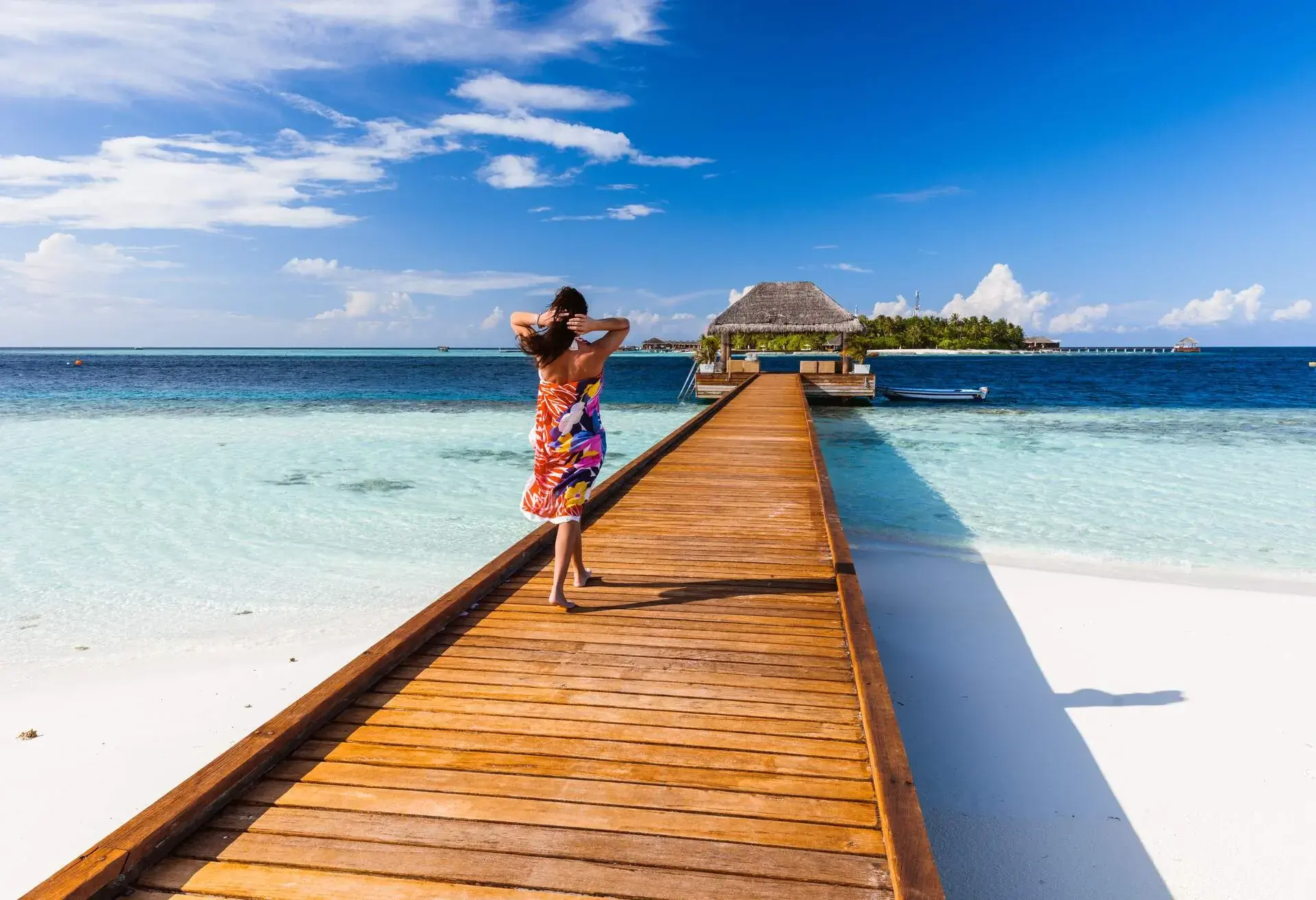 A woman in a colourful sarong walks on a wooden boardwalk across an azure beach that leads to an overwater cottage.