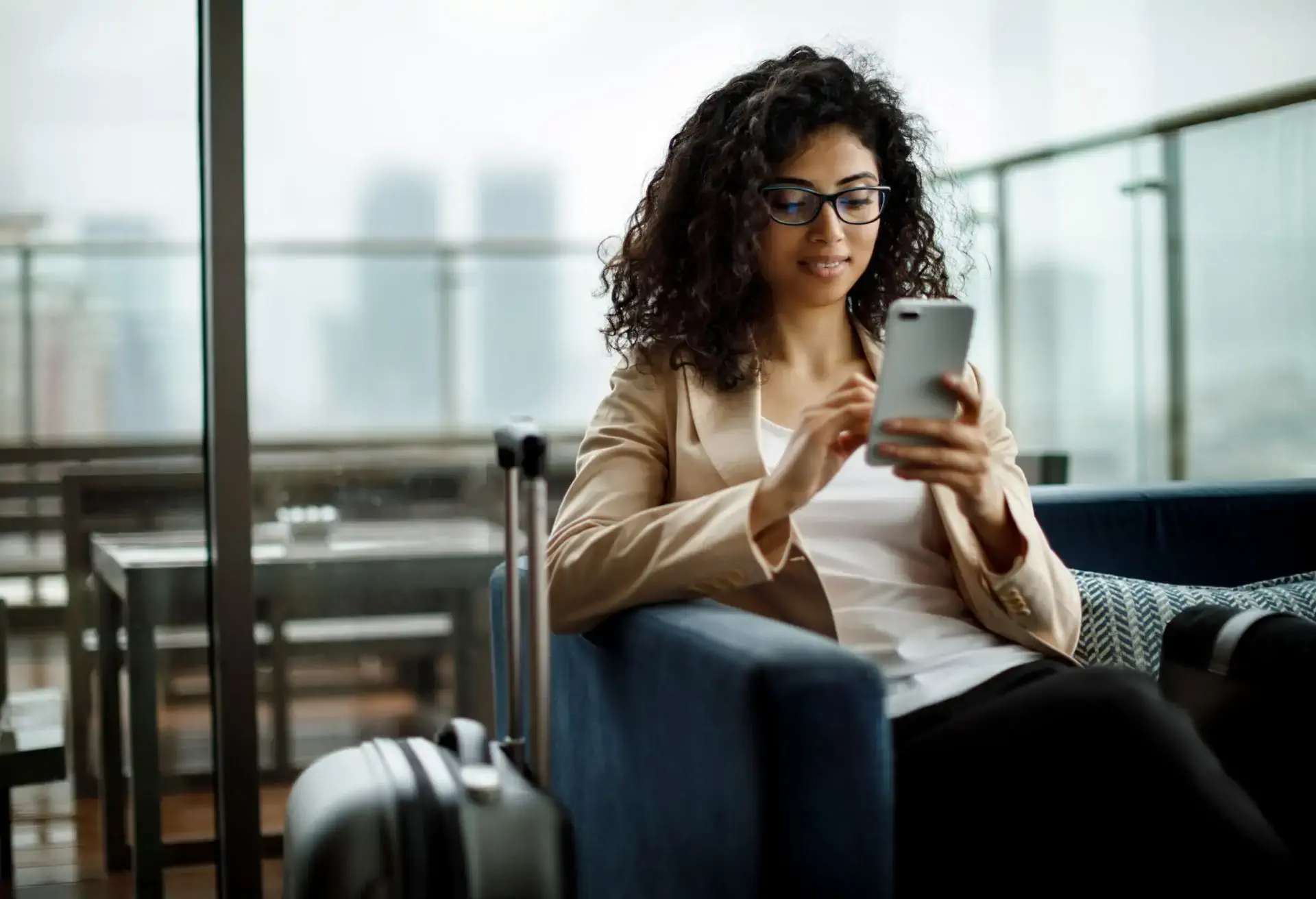 Woman at airport lounge using phone, suitcase beside her