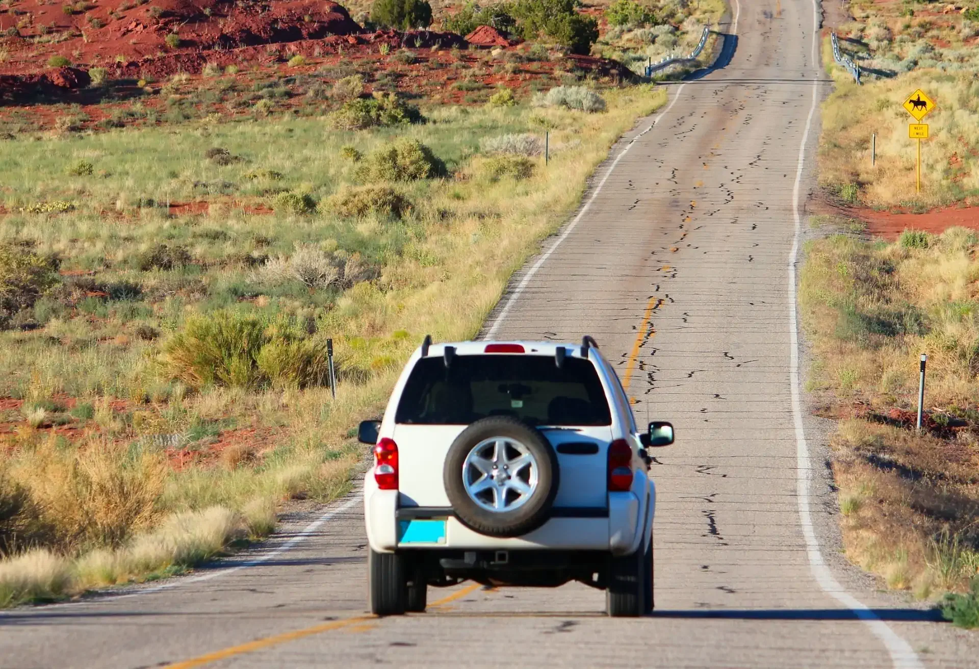 Carro SUV branco circulando por uma estrada longa e deserta rodeada por vegetação rasteira em um cenário ensolarado.