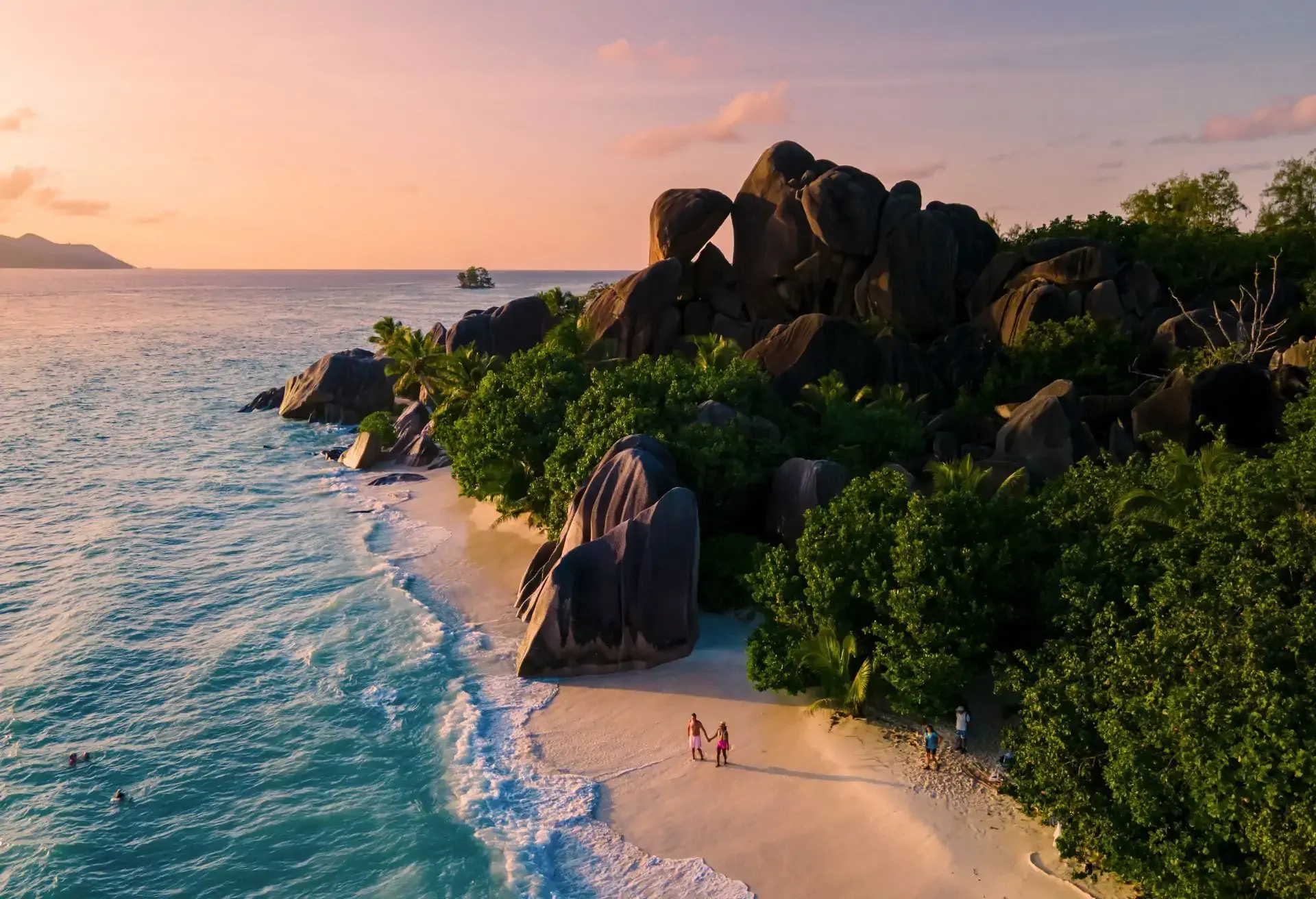 Tropical beach at sunset with turquoise waves, lush greenery, and giant granite boulders on the shore.
