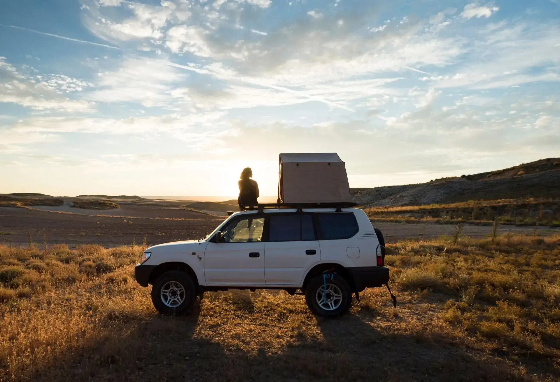 Carro 4x4 com barraca de teto estacionado em campo aberto, com uma pessoa apreciando o pôr do sol ao fundo.