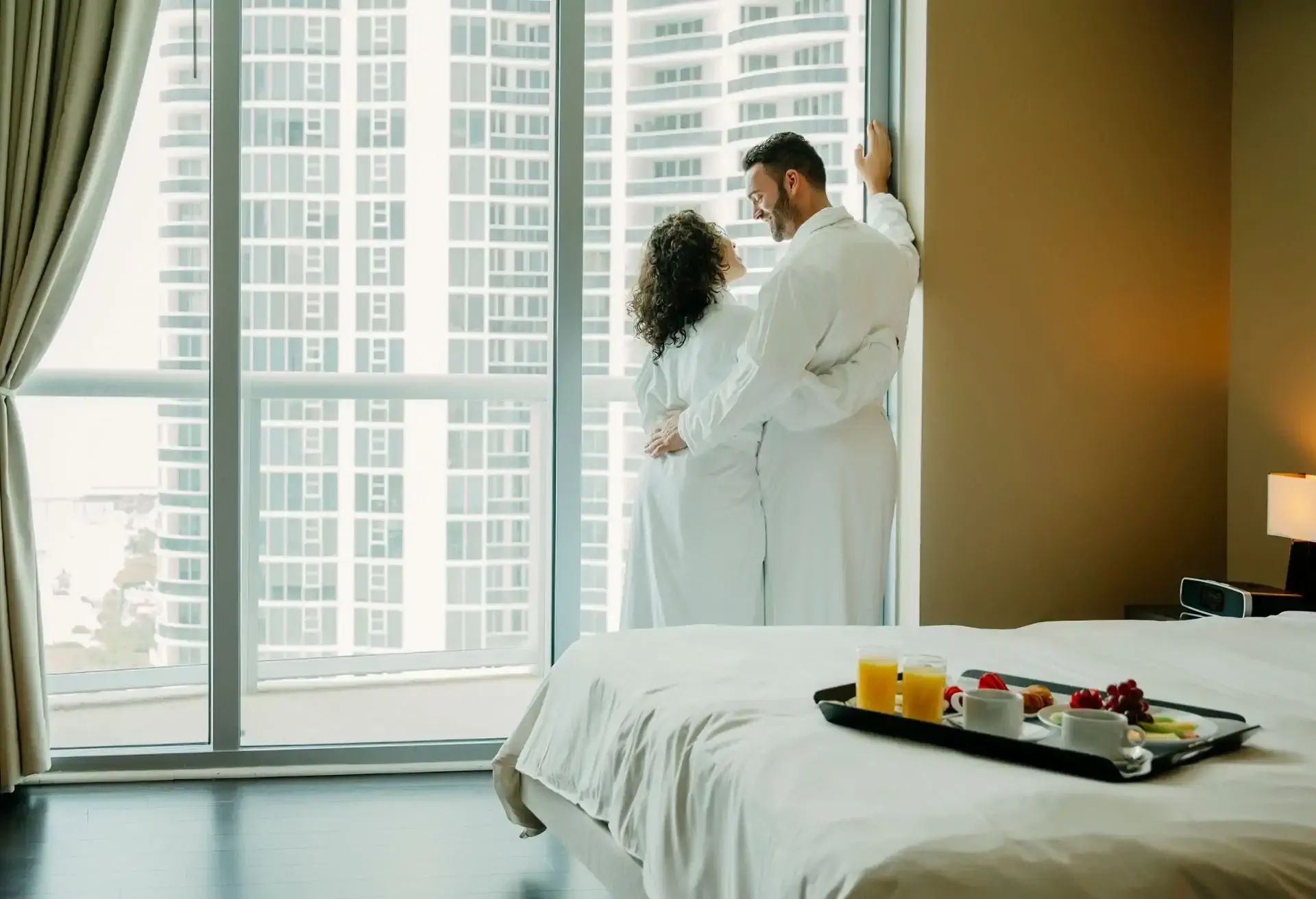 Casal de roupão se abraçando em frente à janela de um quarto de hotel com vista para edifícios altos da cidade.