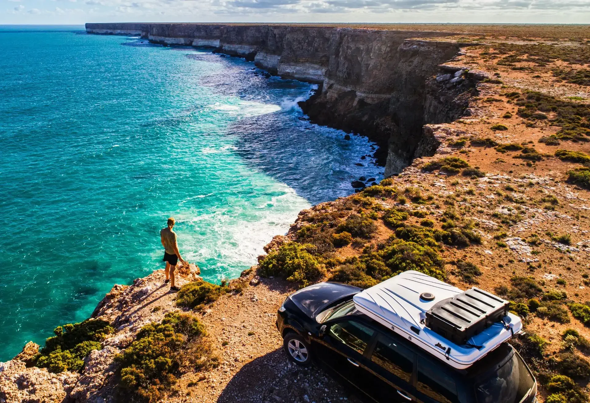 Veículo estacionado à beira de um penhasco com vista panorâmica para o mar azul e uma pessoa observando a paisagem.