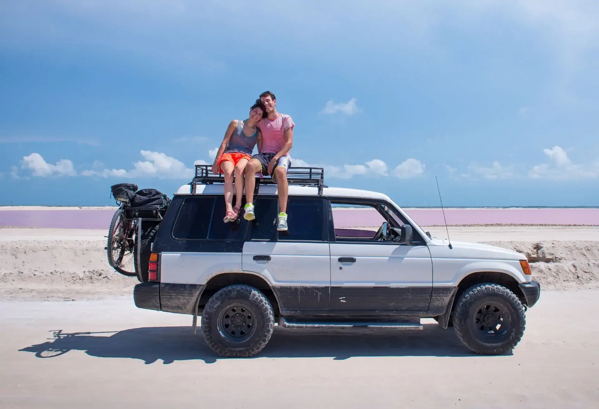 Casal sentado sorridente no teto de um carro SUV estacionado, com céu azul ao fundo e clima descontraído de férias.