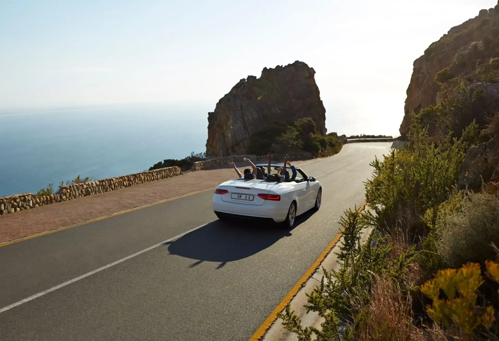 Carro conversível branco circulando por uma estrada costeira com vista para o mar e rochas ao entardecer.