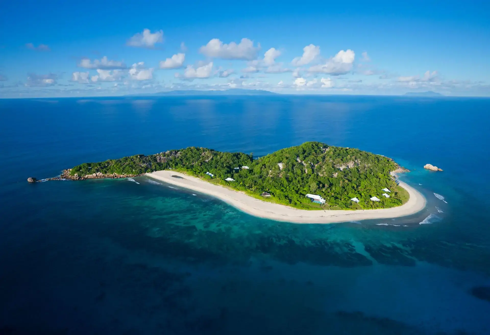 Vista aérea de uma ilha tropical cercada por mar azul e vegetação exuberante, com poucas construções visíveis.