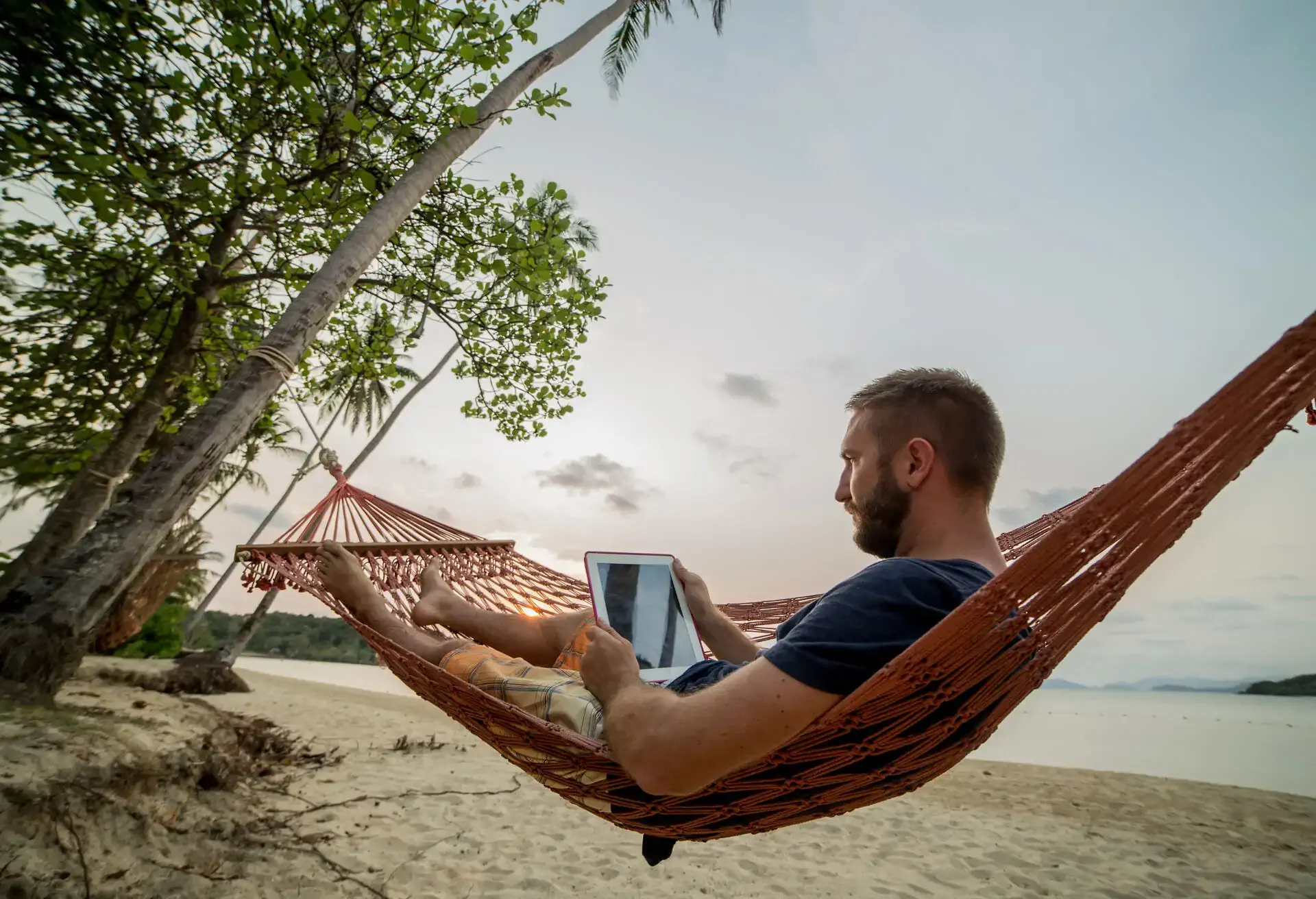 Homem deitado em rede entre árvores na areia, usando um tablet enquanto aprecia o fim de tarde em cenário tranquilo.