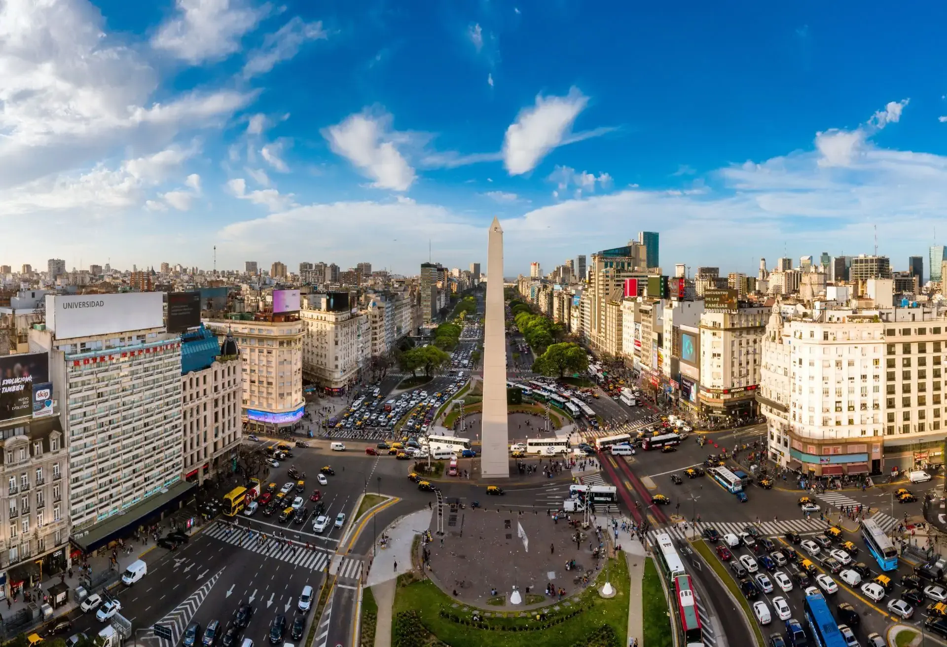 Vista aérea do Obelisco de Buenos Aires com tráfego intenso ao redor, céu azul e prédios históricos ao fundo.