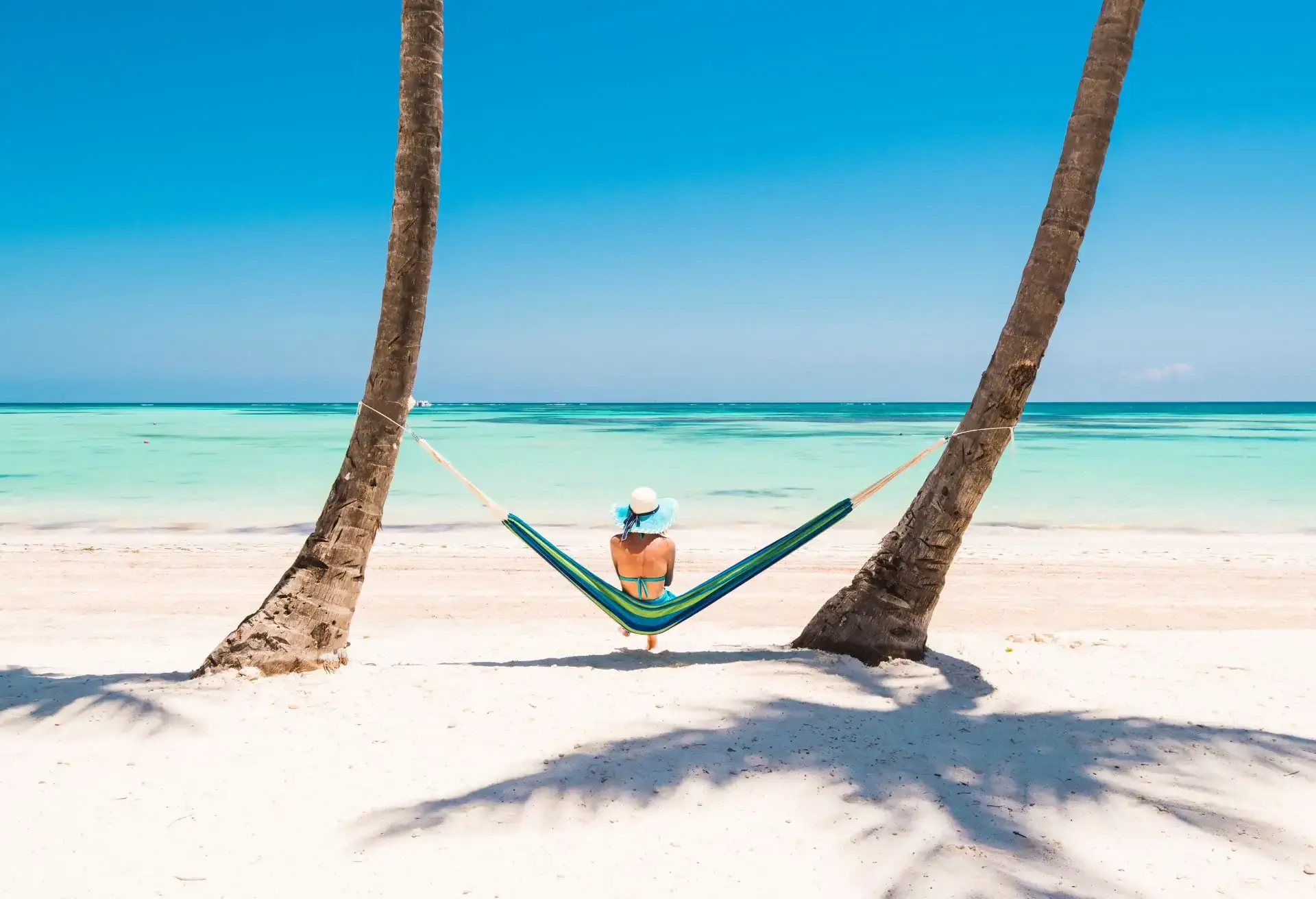 Pessoa relaxando em uma rede esticada entre coqueiros em uma praia de areia branca com mar turquesa cristalino.