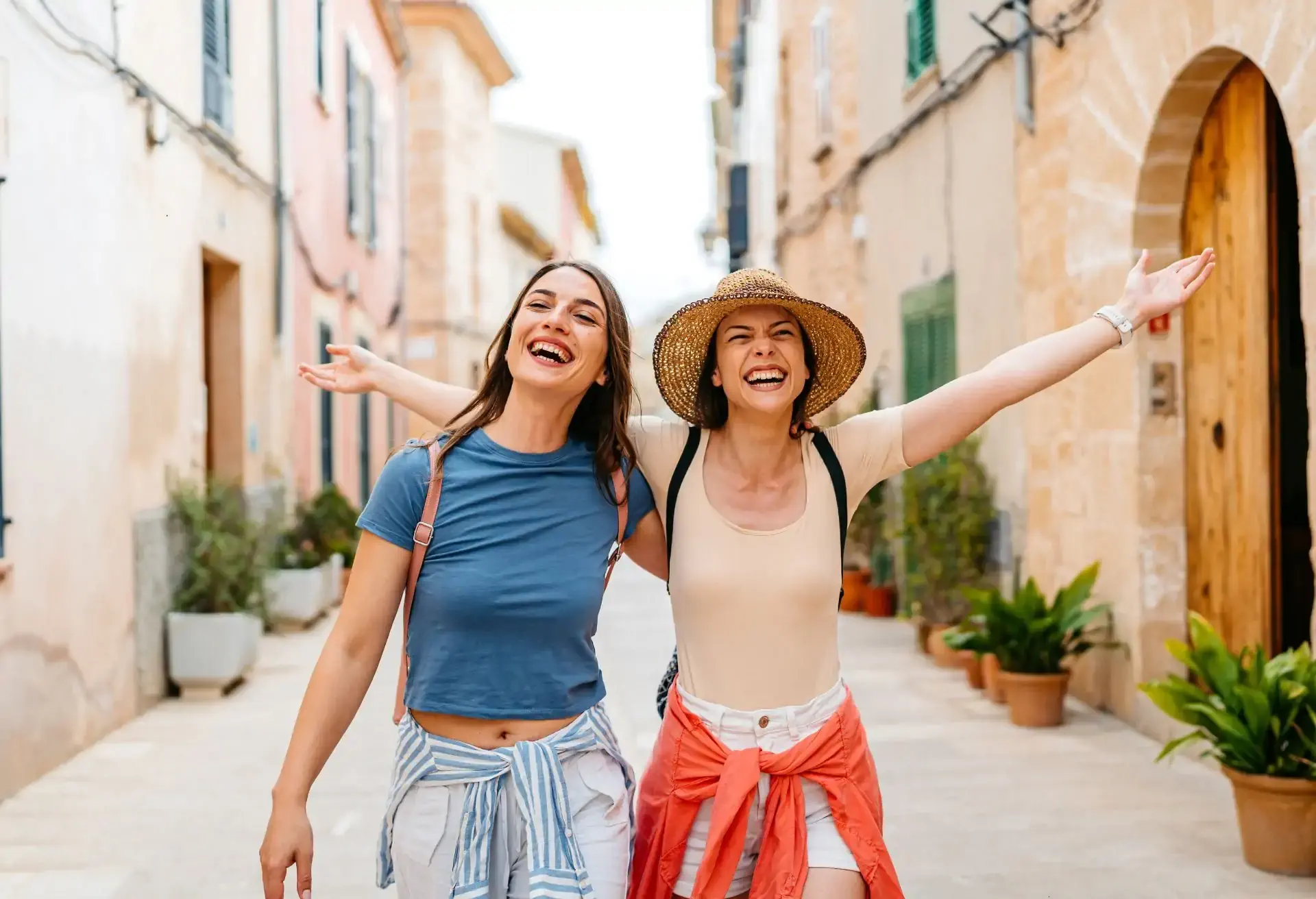 Duas amigas sorrindo com os braços abertos enquanto caminham por uma ruela charmosa com casas coloridas e plantas.