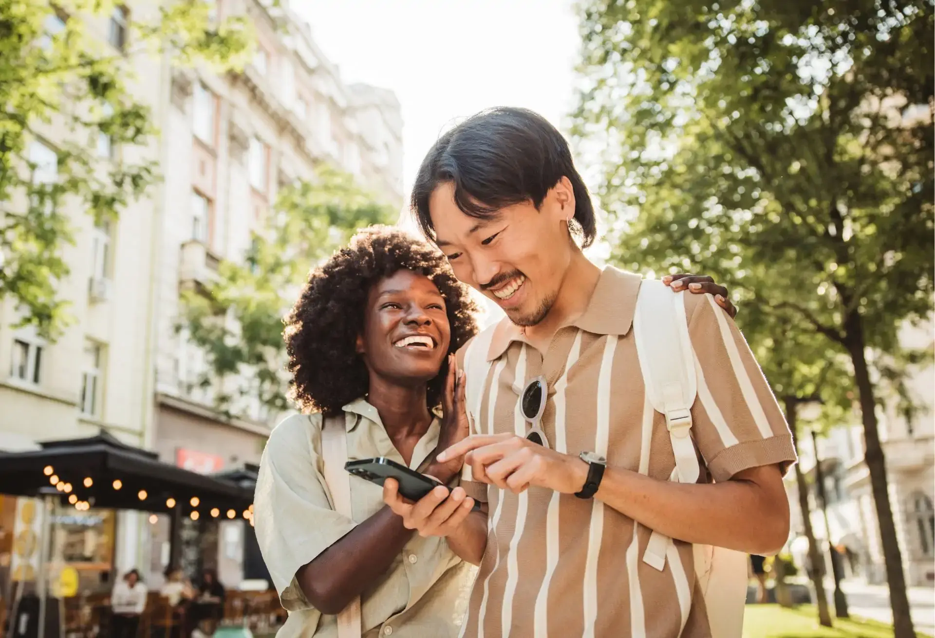 Casal sorridente olhando juntos para um celular em rua arborizada, com mochilas nas costas em ambiente urbano ensolarado.